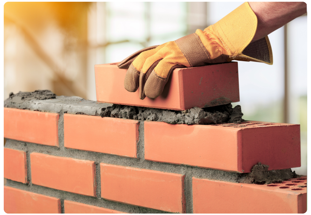 Hand wearing a work glove placing a red brick on wet mortar in a brick wall under construction.