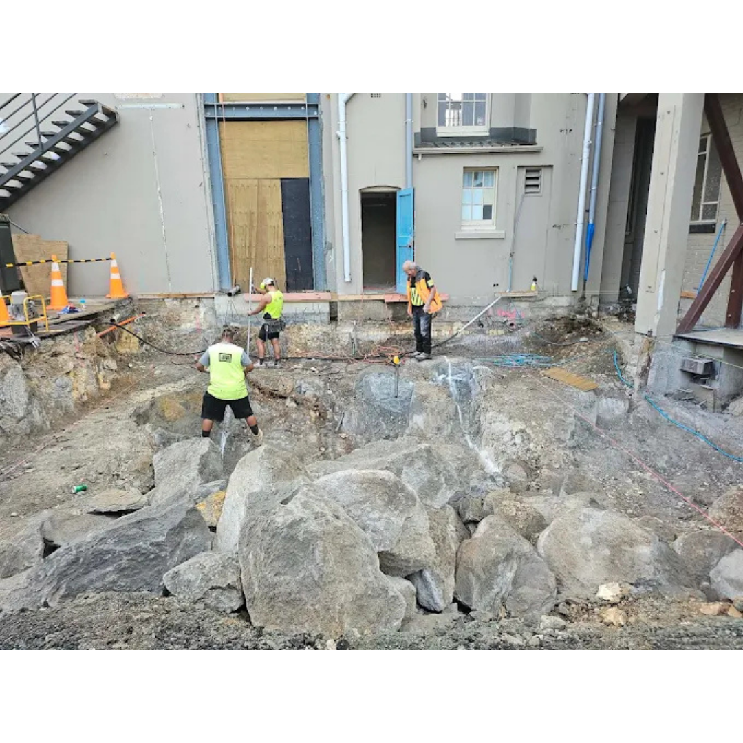 Three construction workers with safety vests working in a deep rocky excavation site next to a building.