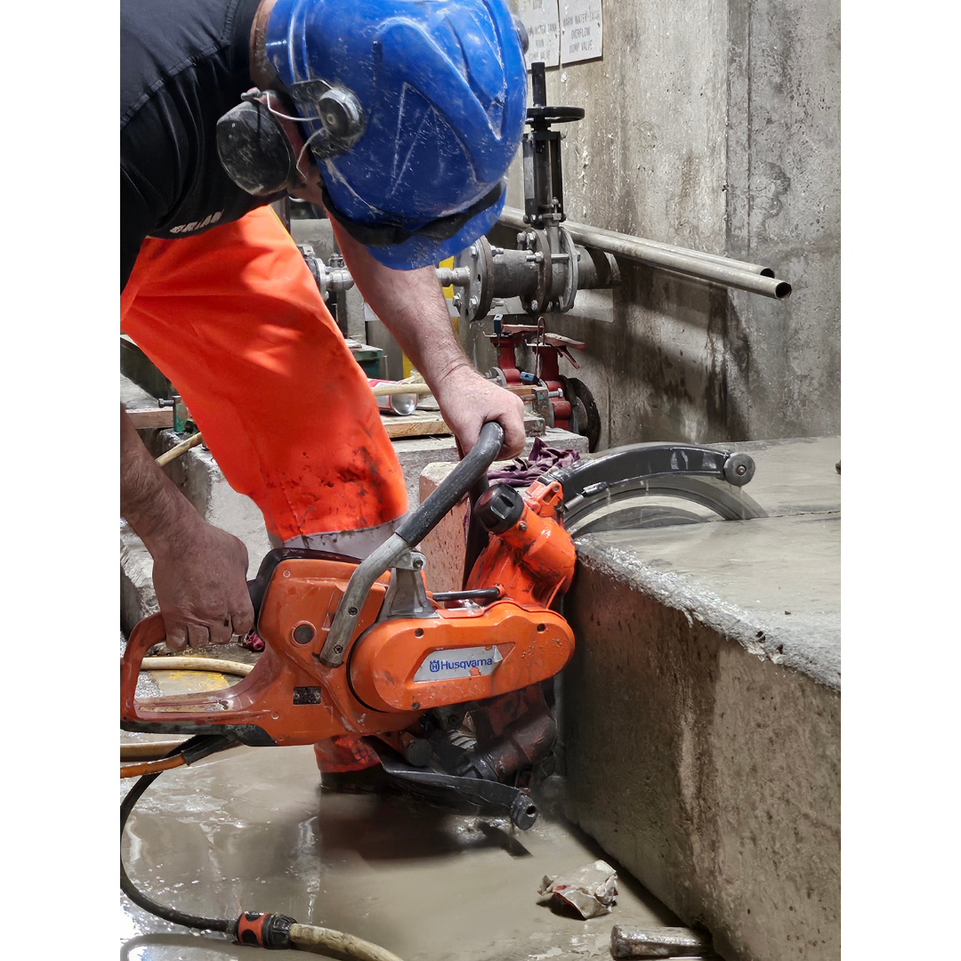 Construction worker wearing blue helmet and orange pants using a Husqvarna saw to cut concrete.