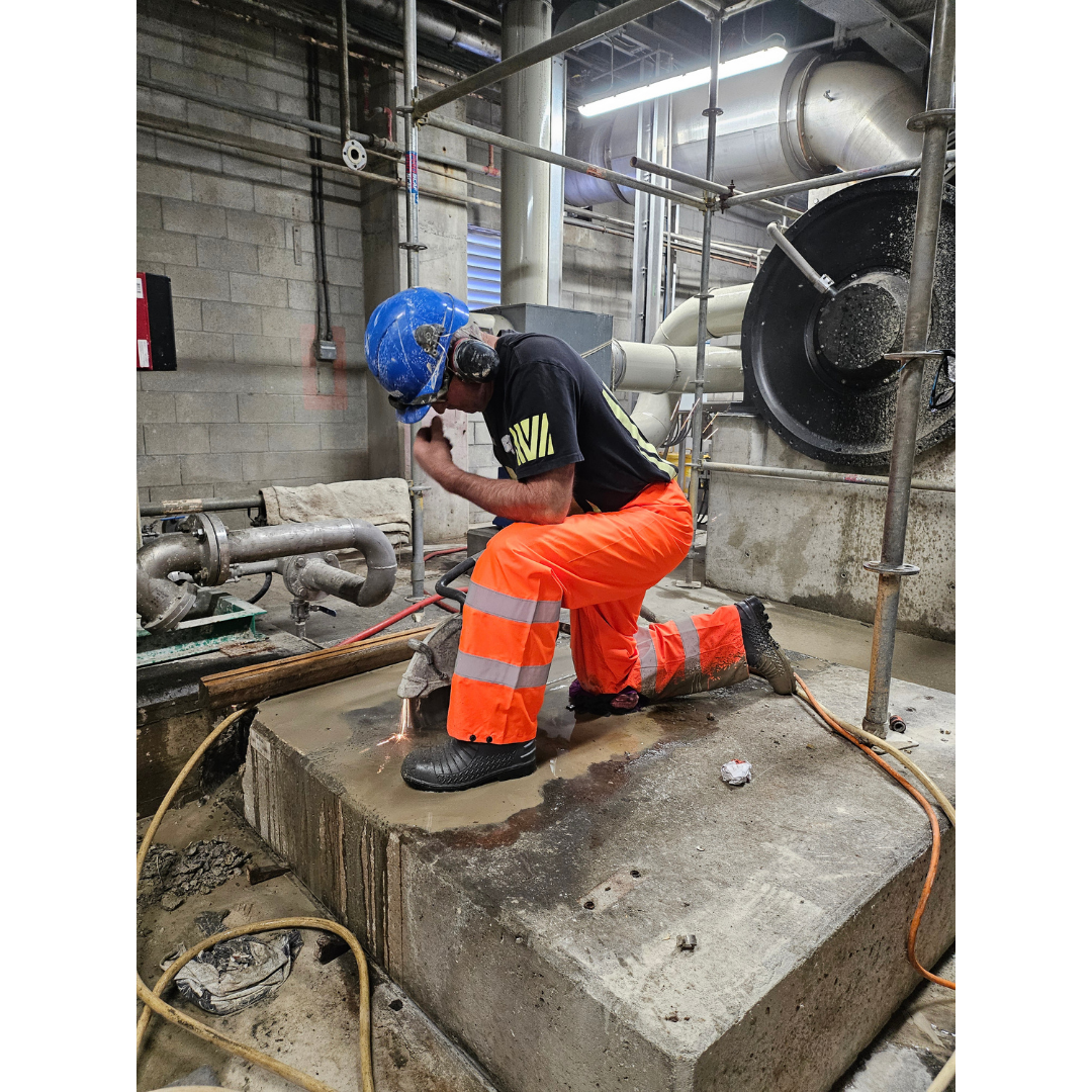 Construction worker in orange reflective pants and blue helmet cutting concrete with a power saw indoors.