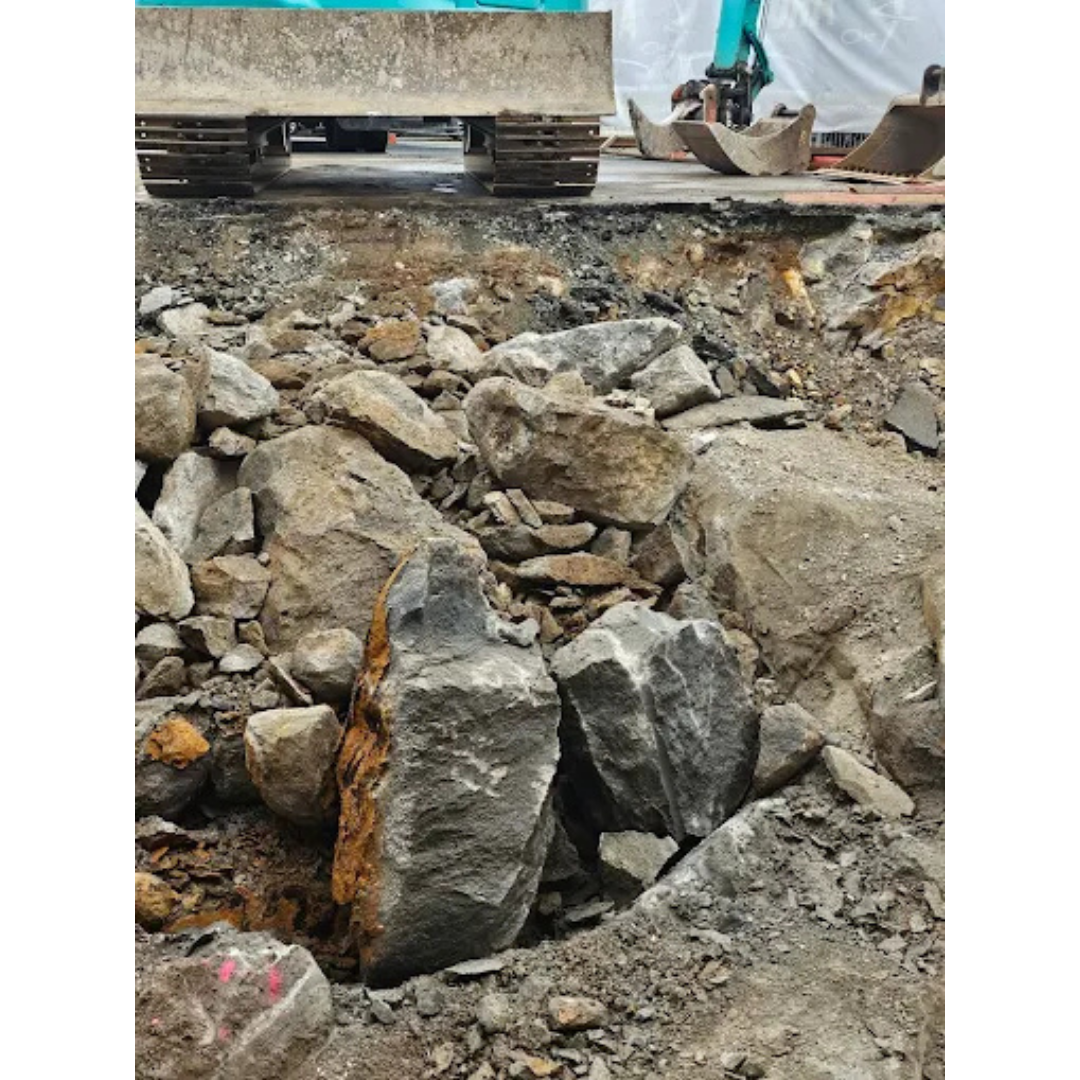 Construction site with excavated ground filled with large broken rocks and heavy machinery tracks visible above.