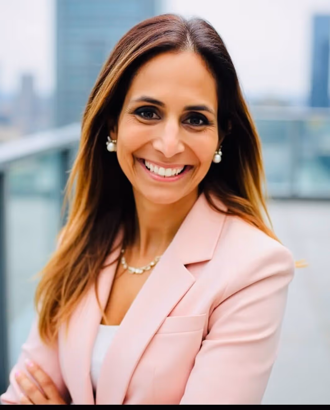 Smiling woman with long brown hair wearing pearl earrings, a white blouse, and a navy blazer.