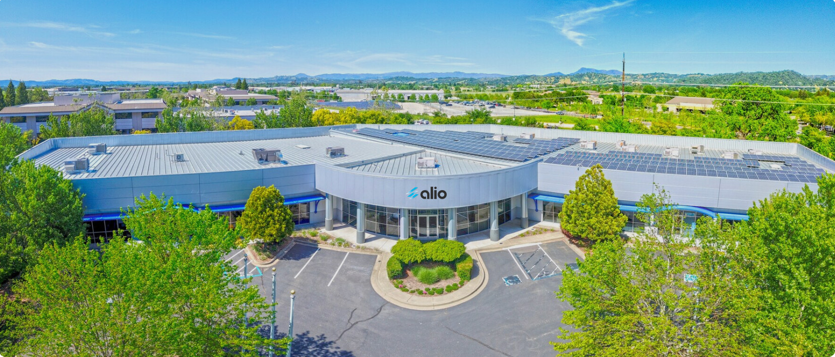 Aerial view of the Alio Labs facility in Santa Rosa, California — a modern single-story industrial building with the Alio logo above the main entrance, a large solar panel array covering the roof, and the Sonoma County hills visible in the background.