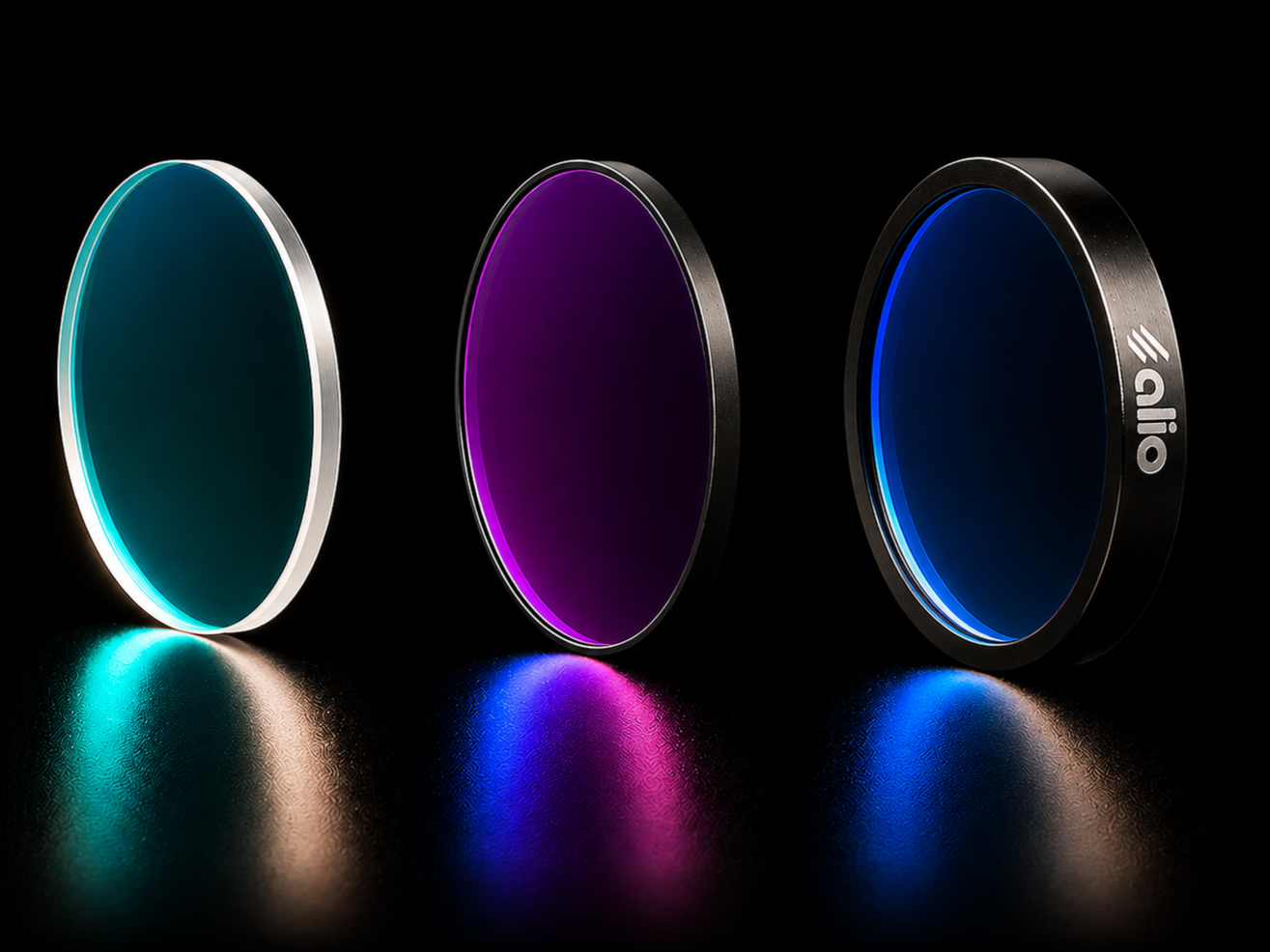 Three circular optical filters standing upright on a reflective surface: an unmounted filter on the left with a teal interference coating, a painted-edge filter in the center with a deep magenta and purple coating, and a fully mounted filter on the right with a deep blue coating in a black anodized mount engraved with the Alio Labs logo.