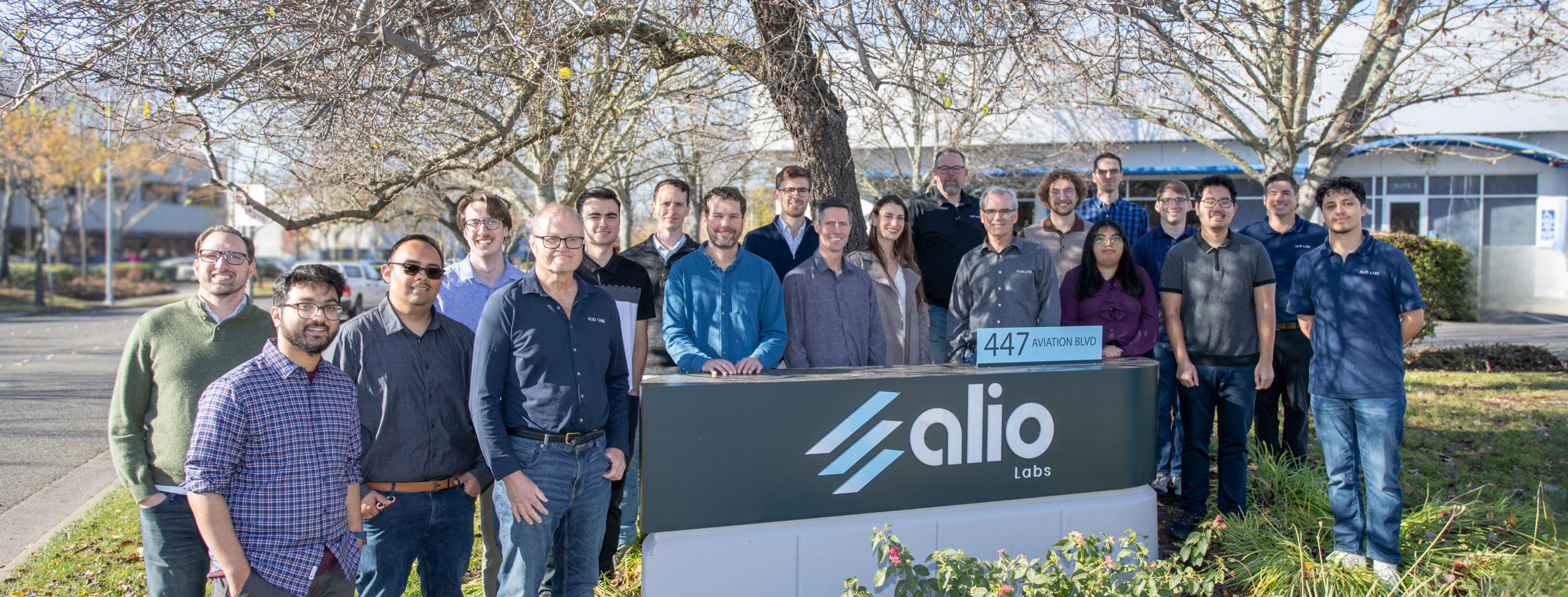 The Alio Labs team gathered outdoors in front of the company sign at 447 Aviation Blvd, Santa Rosa, California, surrounded by trees and flowering plants on a sunny day.