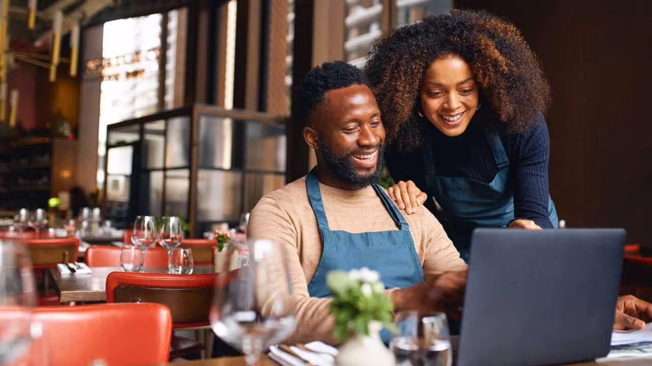 Smiling man and woman wearing aprons looking at a laptop in a restaurant.