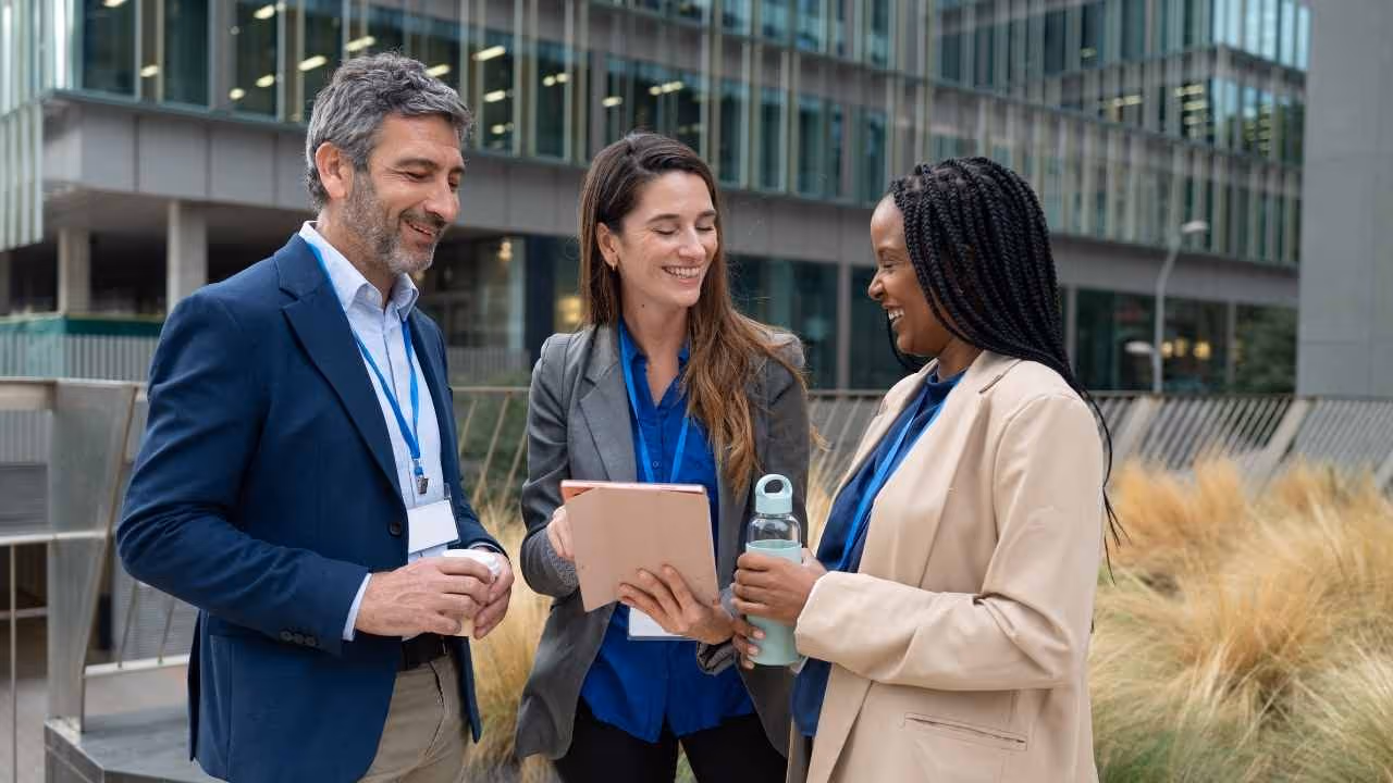 Three business professionals smiling and discussing work outdoors near modern office buildings, with one holding a tablet.