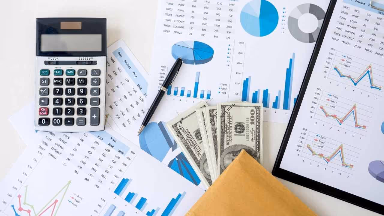 Calculator, pen, printed financial charts, and US dollar bills partially inside a brown envelope on a white surface.
