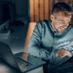 Hombre sonriendo mientras sostiene una tablet frente a una computadora portátil en un ambiente interior con luz tenue.