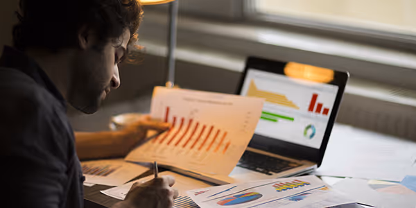 Hombre analizando gráficos y datos impresos mientras revisa información en una computadora portátil.