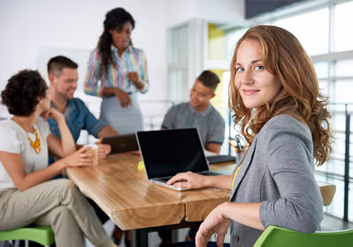 Grupo de cinco personas en una reunión de trabajo alrededor de una mesa con computadora portátil, una mujer sonriente mira a la cámara.