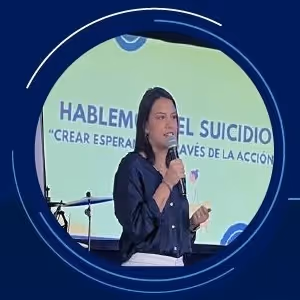Mujer dando una charla sobre prevención del suicidio con un micrófono, frente a una pantalla que dice 'Hablemos del suicidio, crear esperanza a través de la acción'.