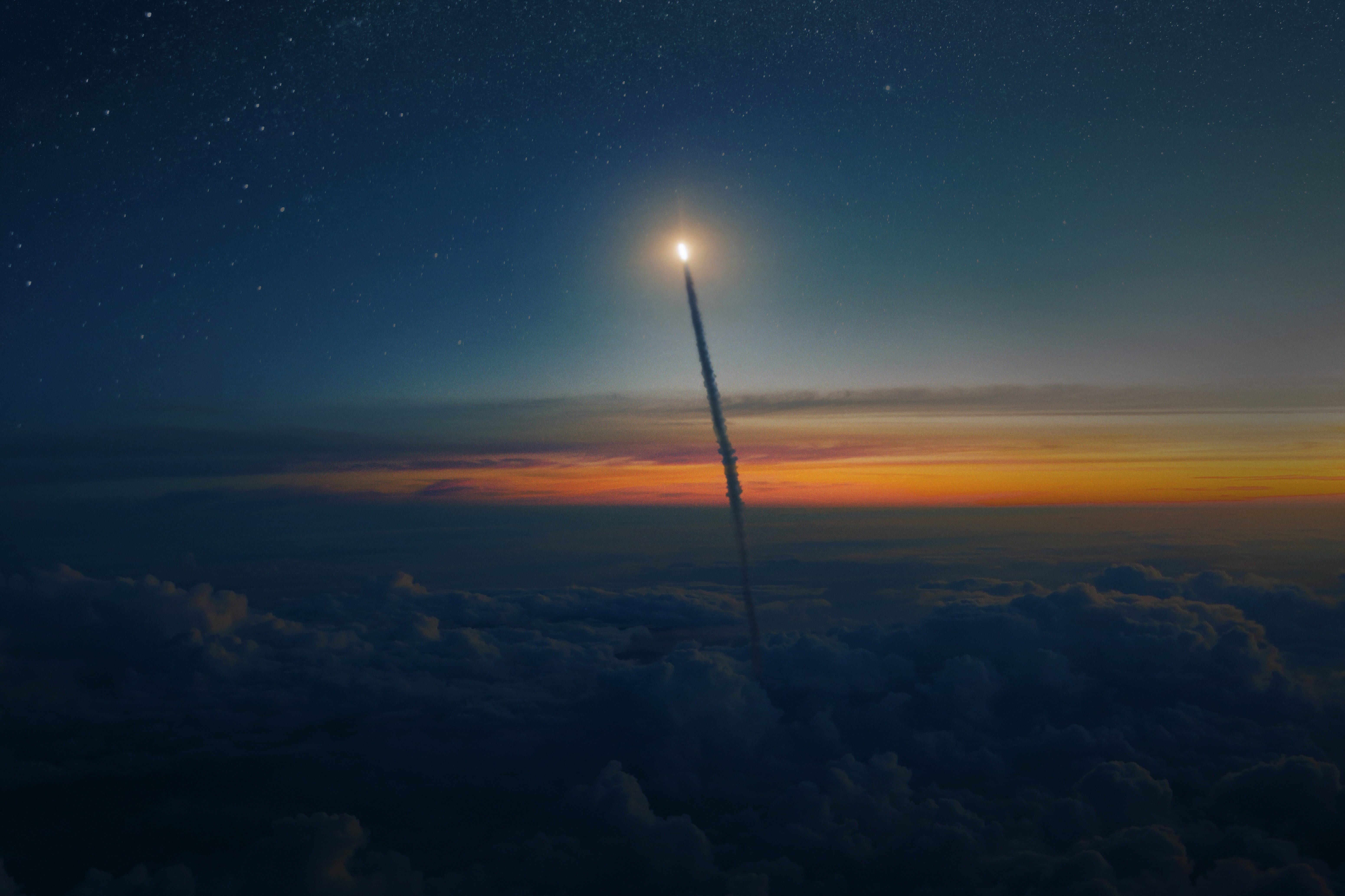 Rocket launching through a starry sky at sunset above dense clouds with a bright exhaust trail.