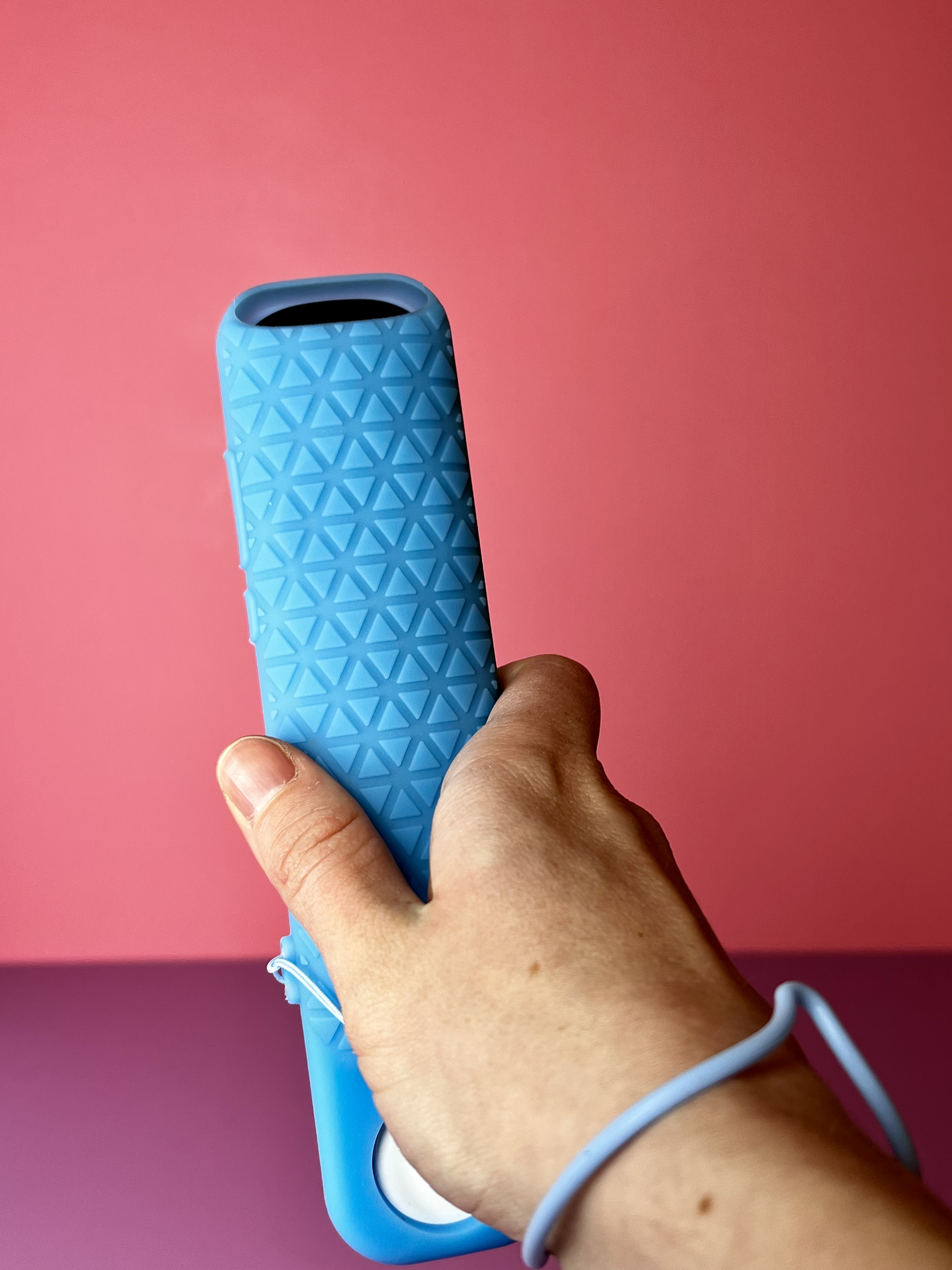 A hand holding the back of a bright blue textured remote case with a wrist strap, shown against a pink background.