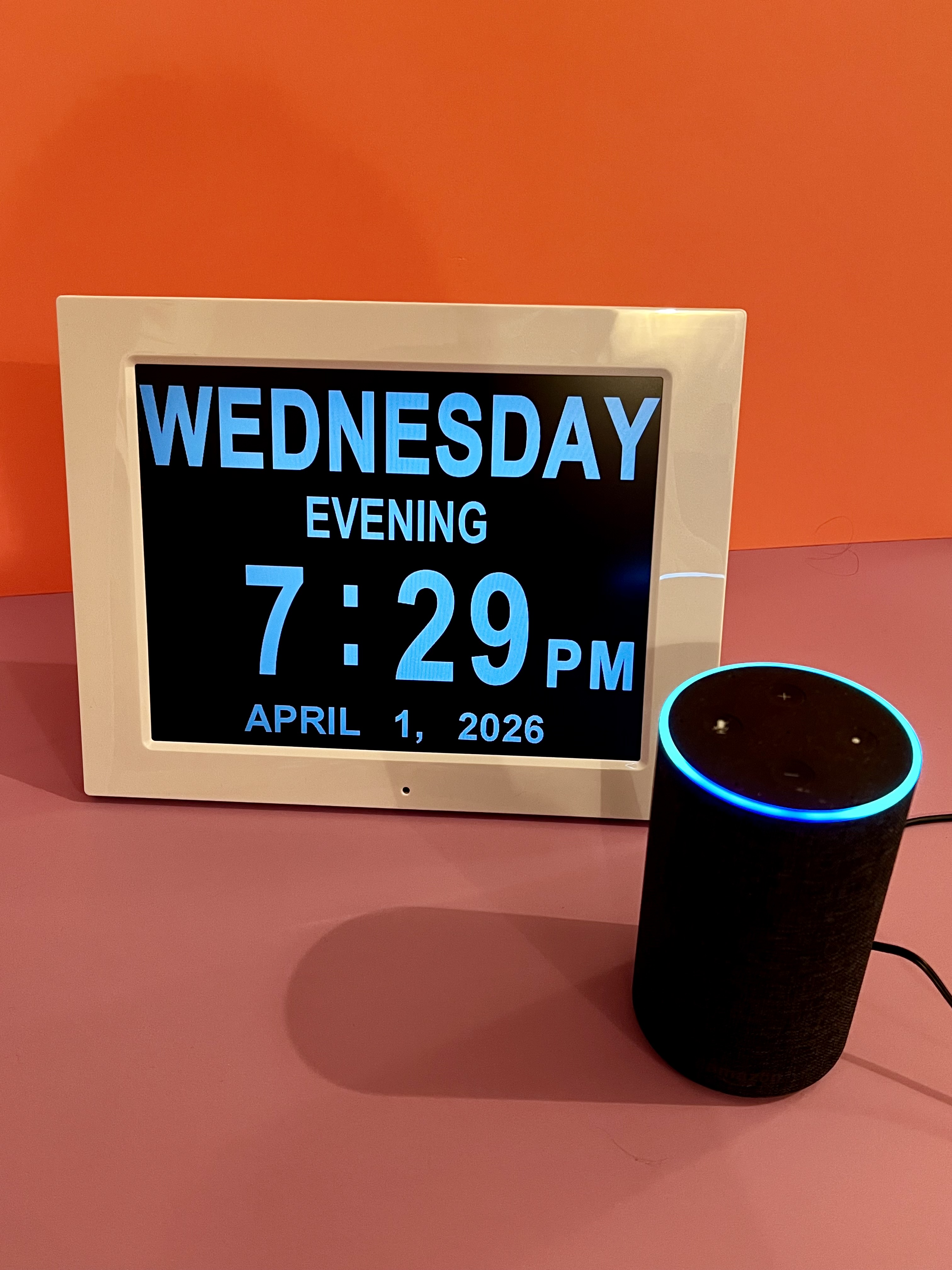 A digital day and time clock displayed beside a black smart speaker on an orange and pink background.