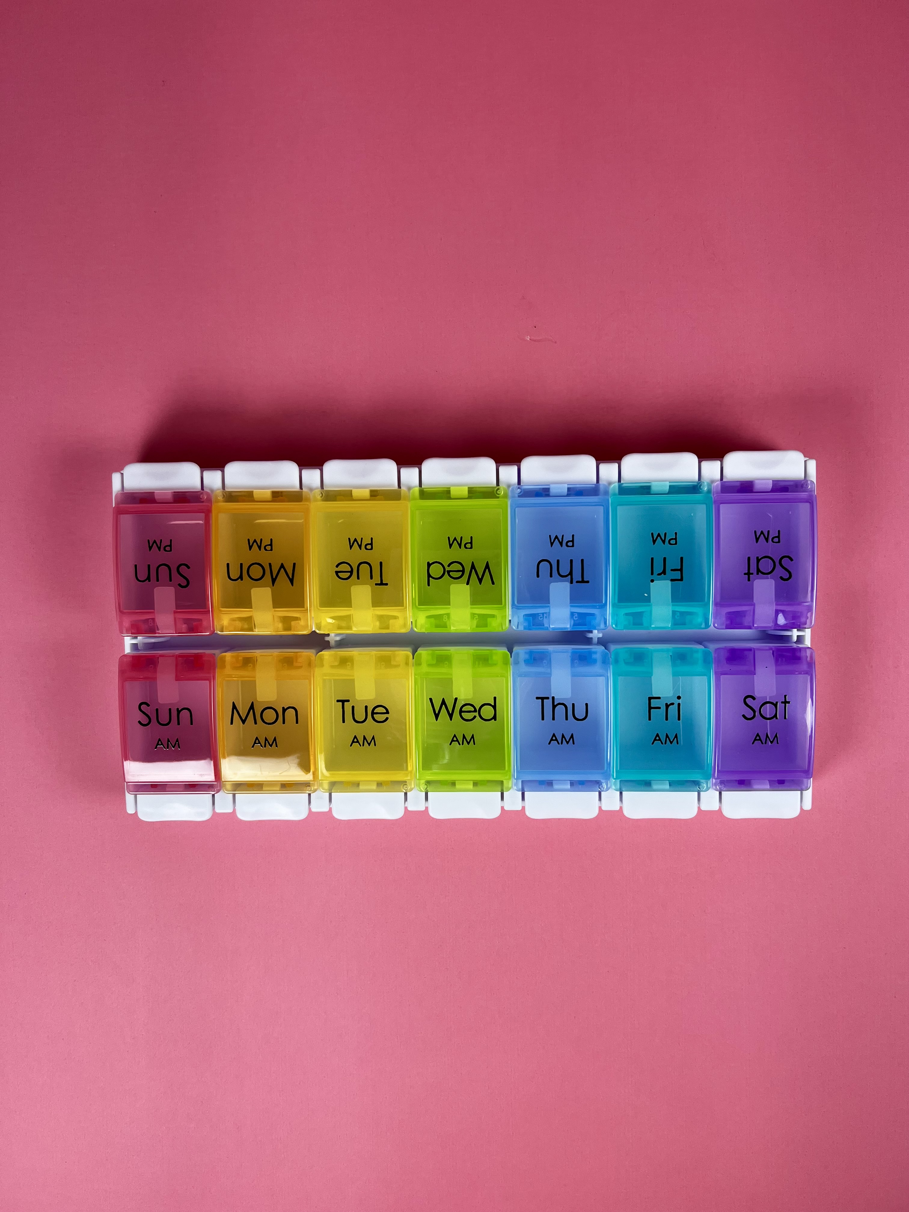 A color-coded pillbox with large day-of-the-week labels on a pink background.