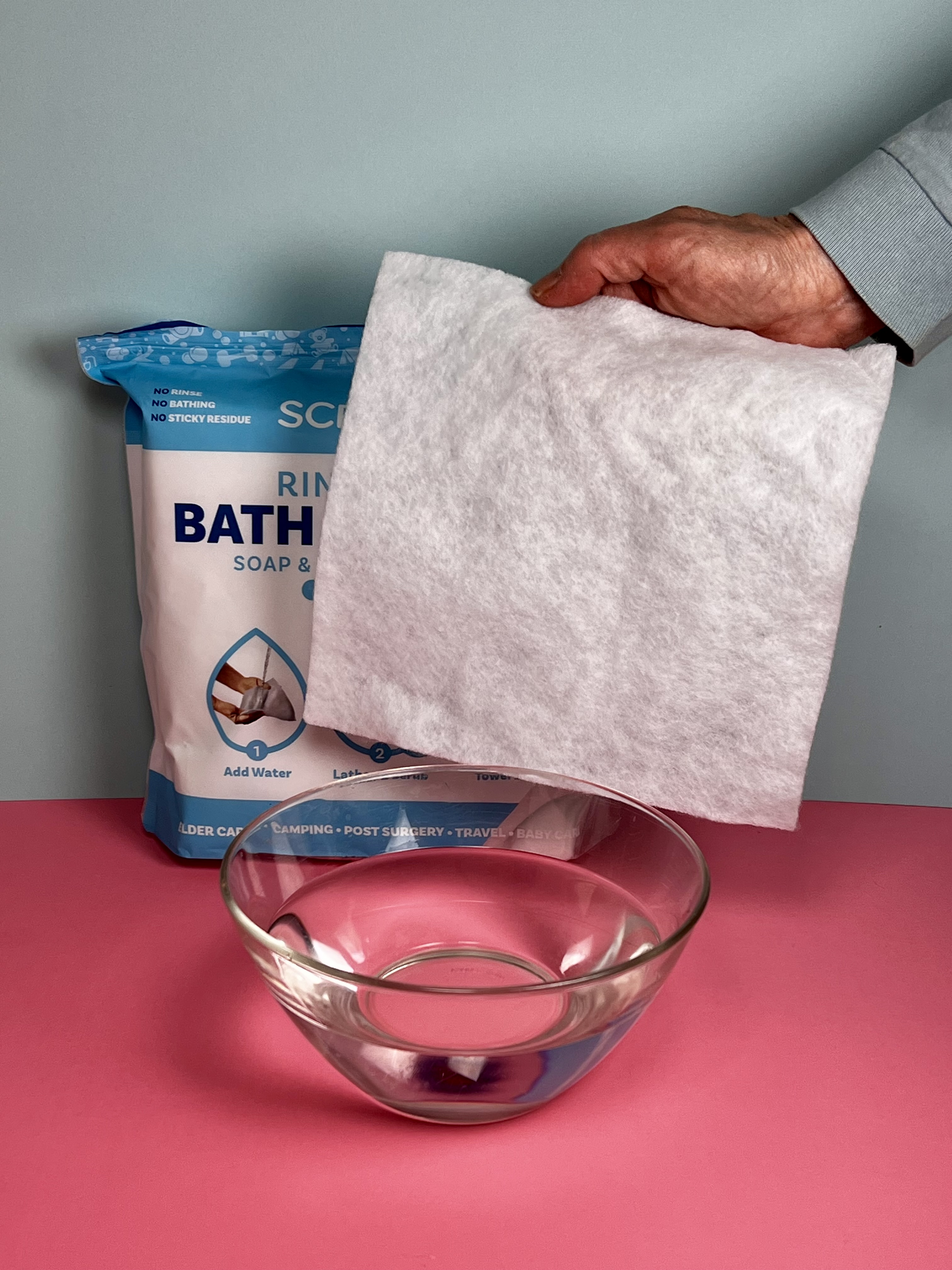 A hand holding a white rinse-free bath sponge sheet beside its blue package and a glass bowl of water.