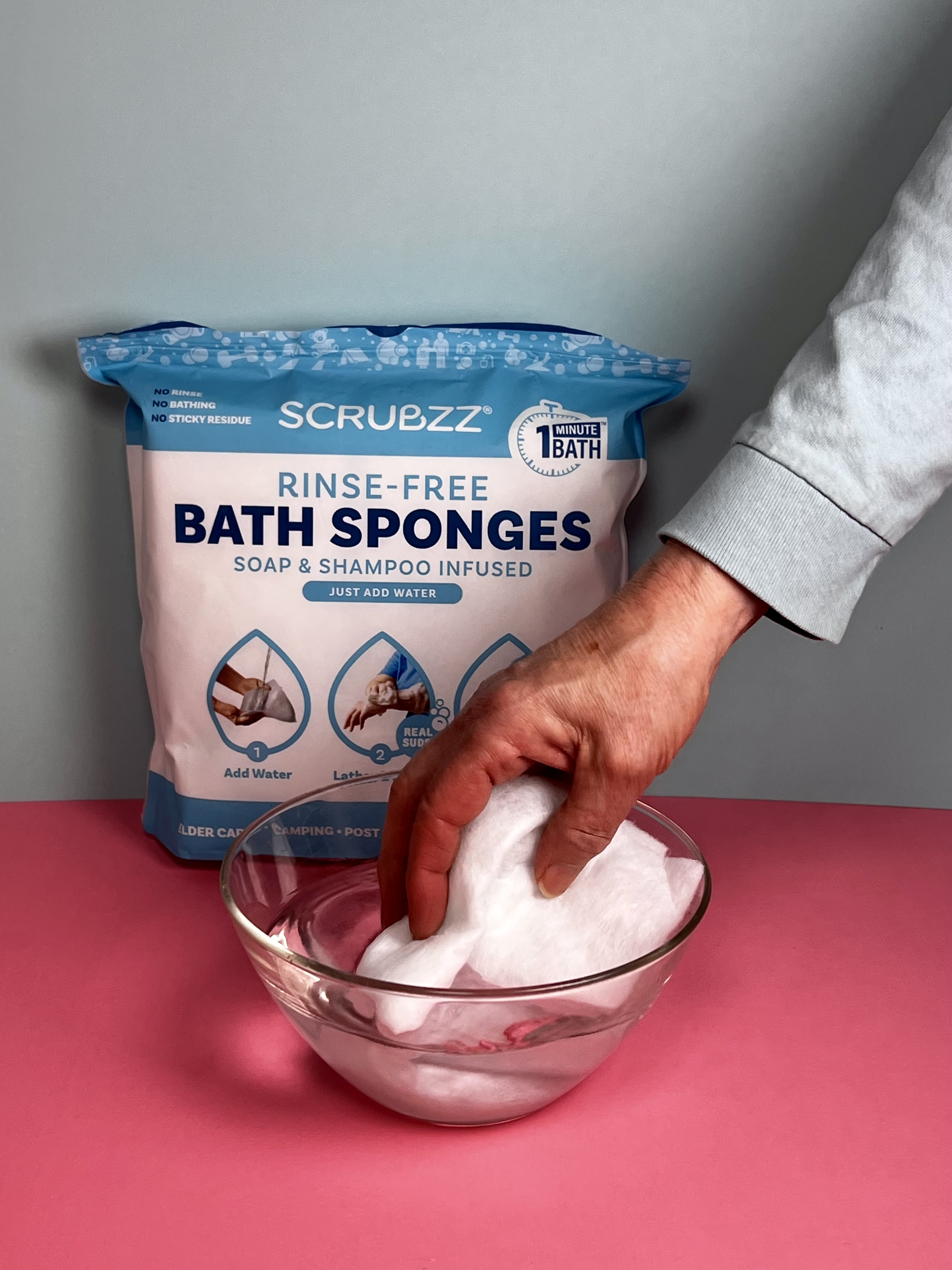 A hand wetting a rinse-free bath sponge sheet in a glass bowl of water, with the product package behind it.