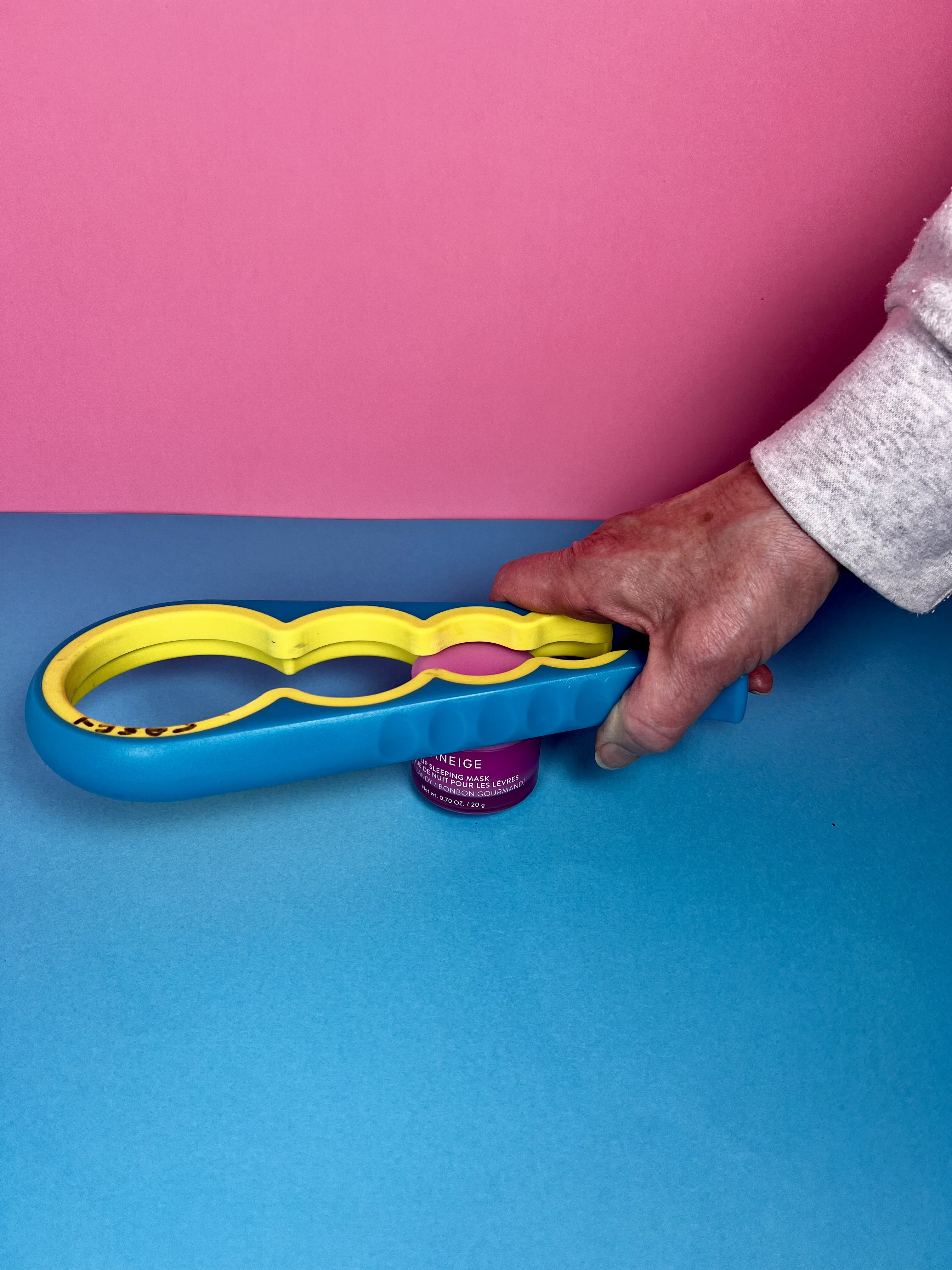 A hand using a colorful multi-size opener to grip and open a small cosmetics jar.