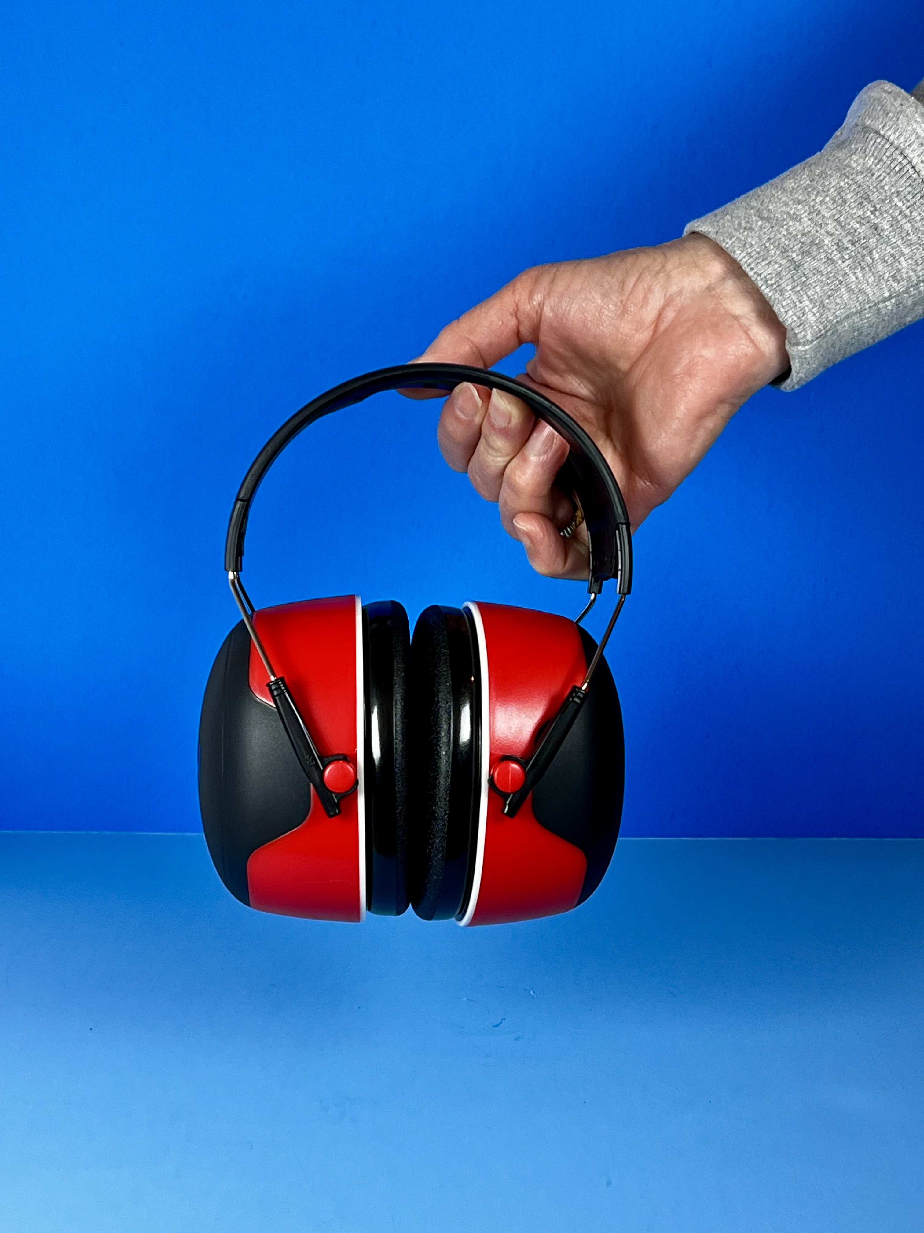 A hand holding red and black noise-reducing earmuffs with cushioned ear cups against a blue background.