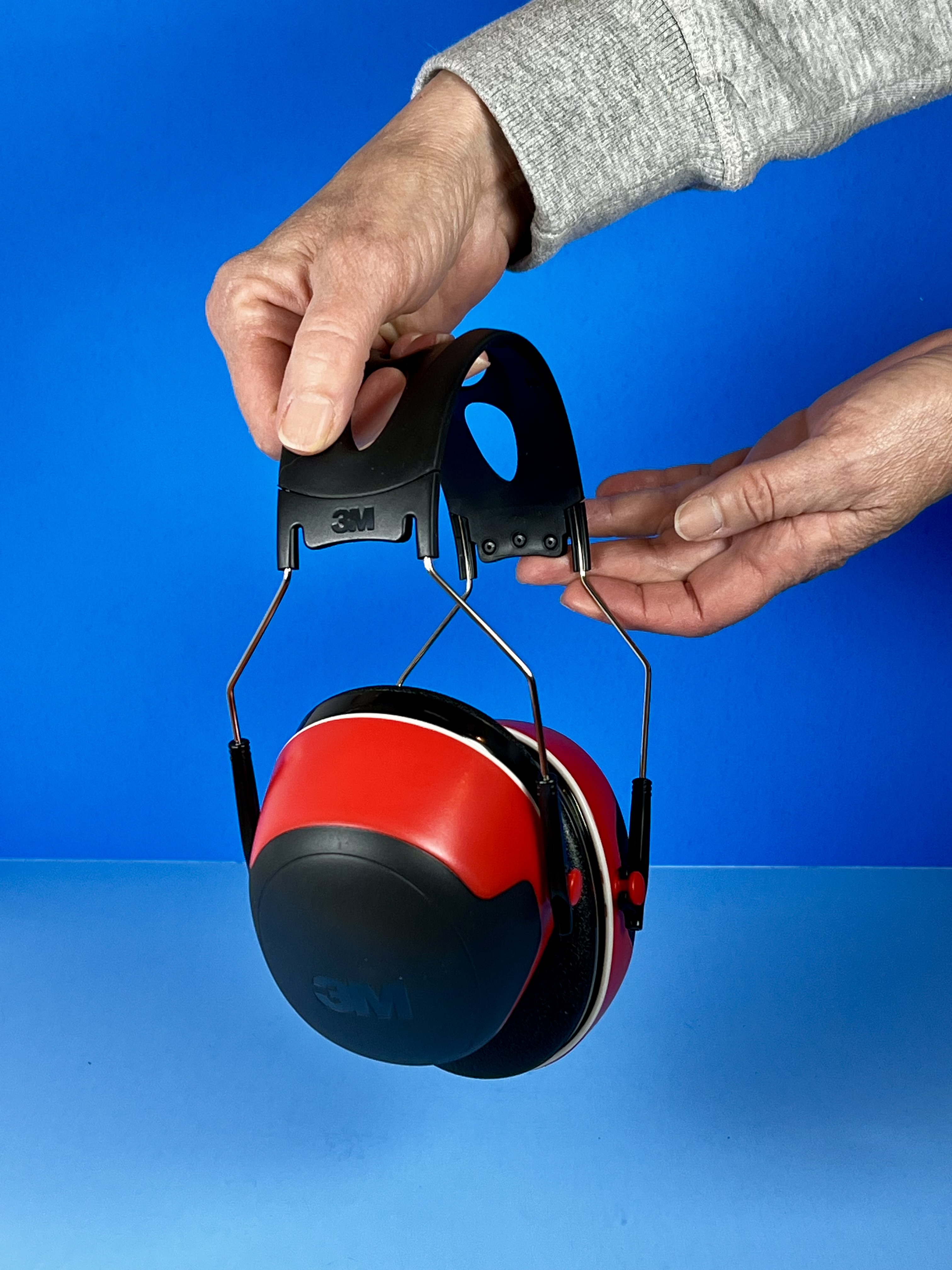 Hands adjusting the headband on red and black 3M noise-reducing earmuffs against a blue background.