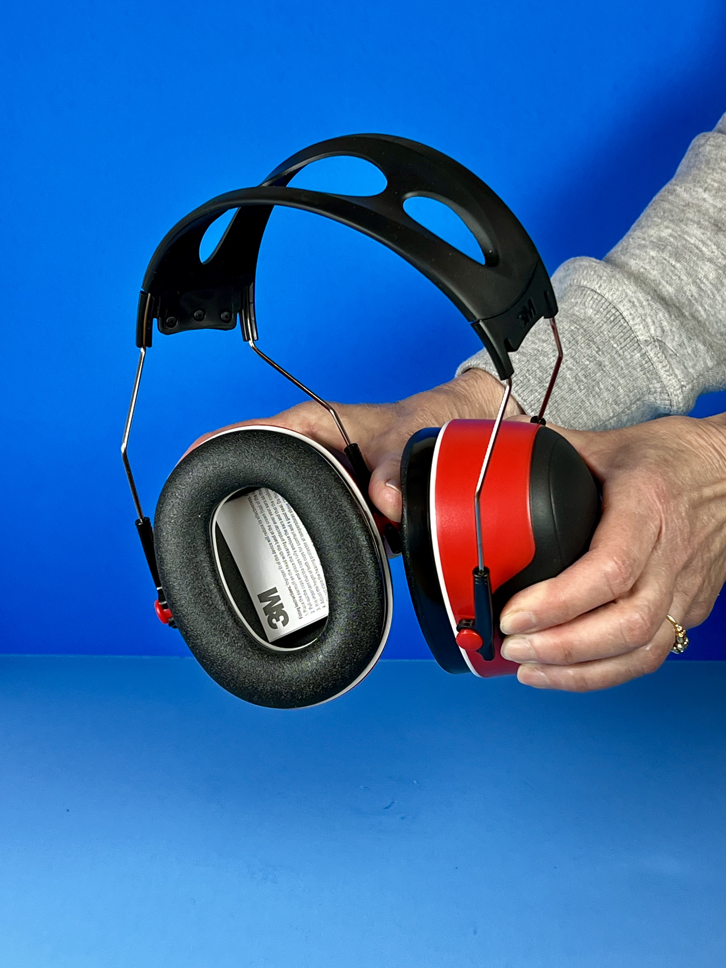 Hands holding red and black 3M noise-reducing earmuffs open to show the cushioned ear cups and adjustable headband against a blue background.