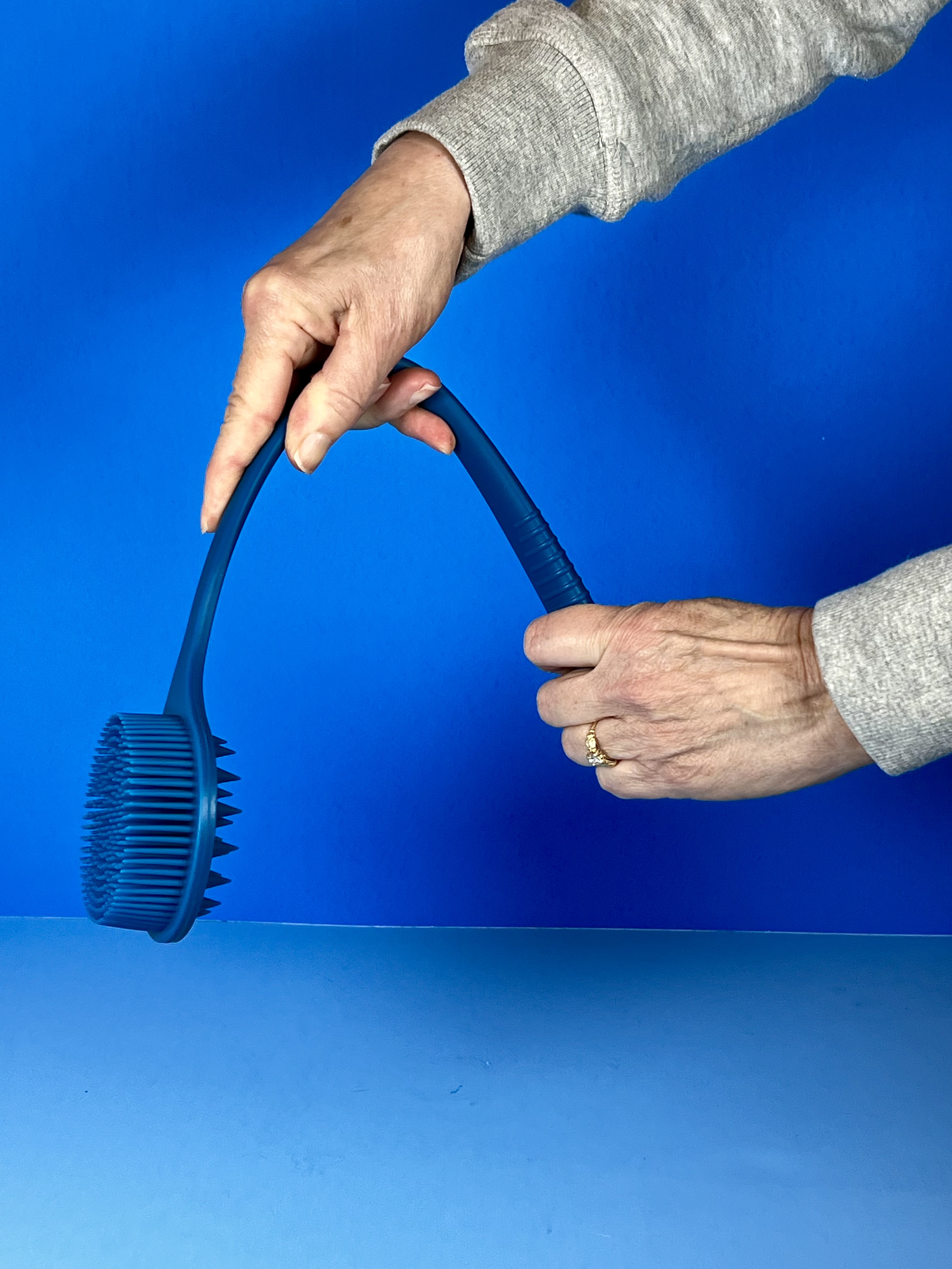 Hands bend a long-handled blue silicone body and hair scrubber, showing its flexible handle and textured scrubber head against a blue background.