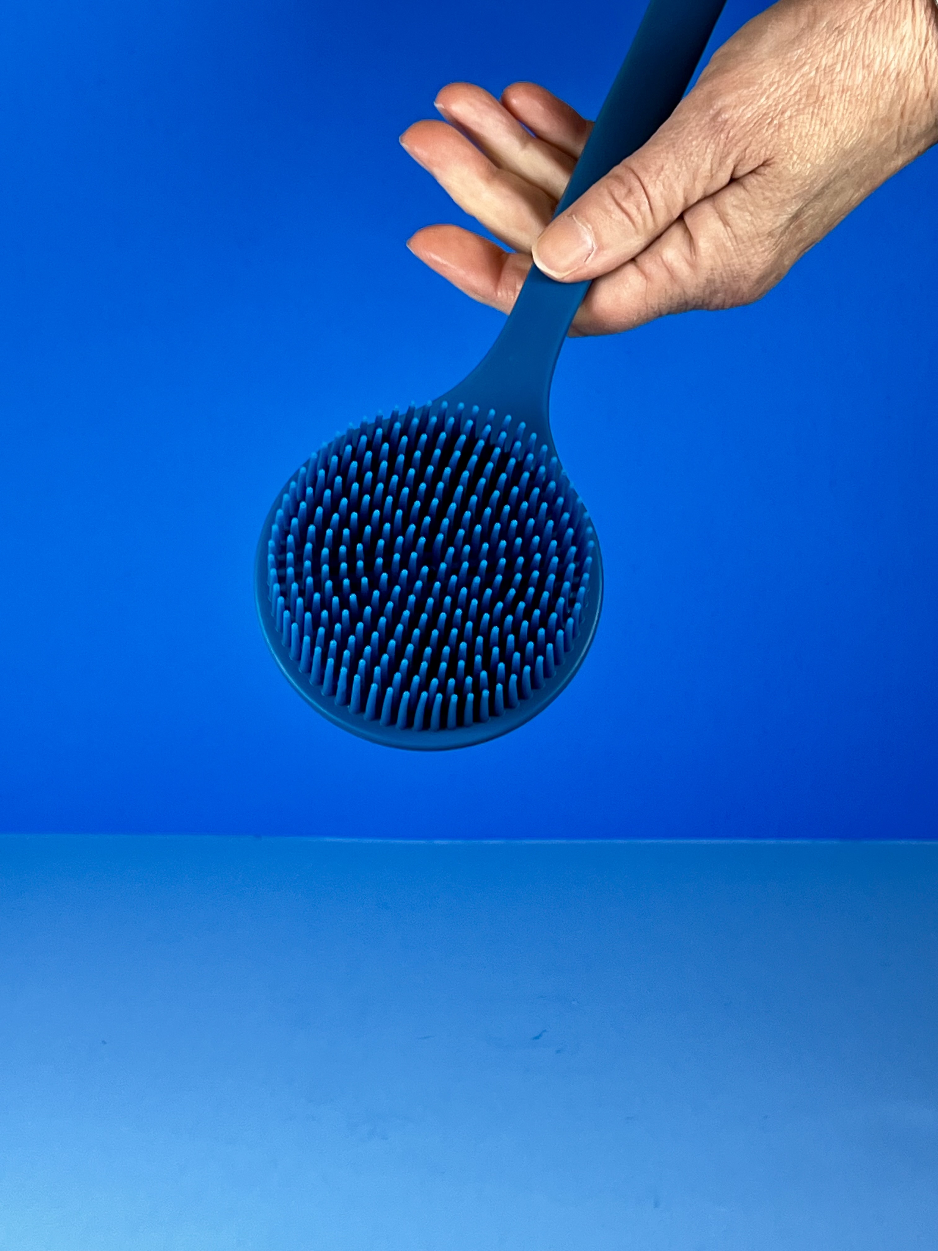 A hand holds a blue long-handled silicone body scrubber, showing the round scrubber head with soft bristles against a blue background.