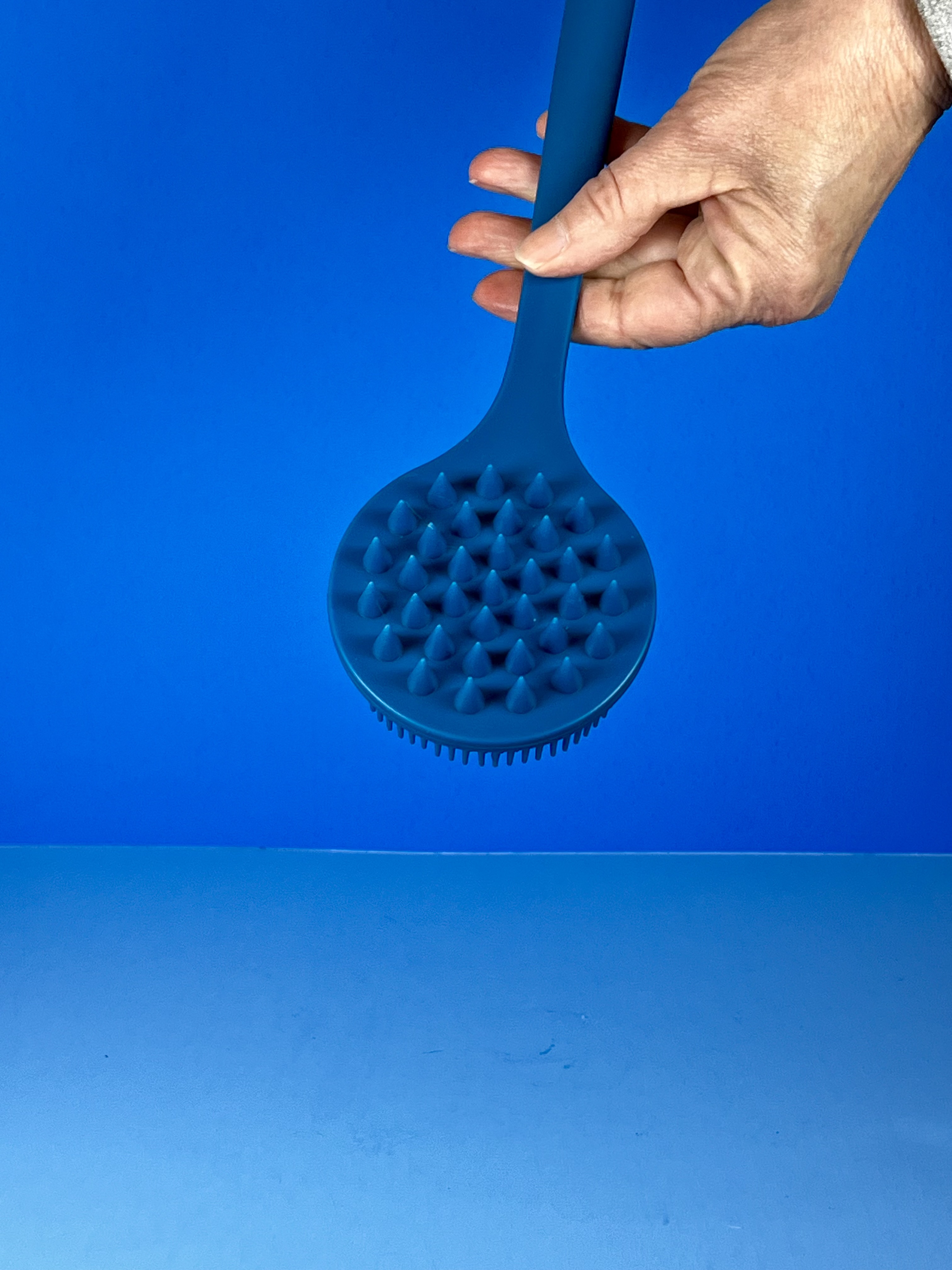 A hand holds the blue long-handled silicone scrubber, showing the round side with raised massage bumps against a blue background.