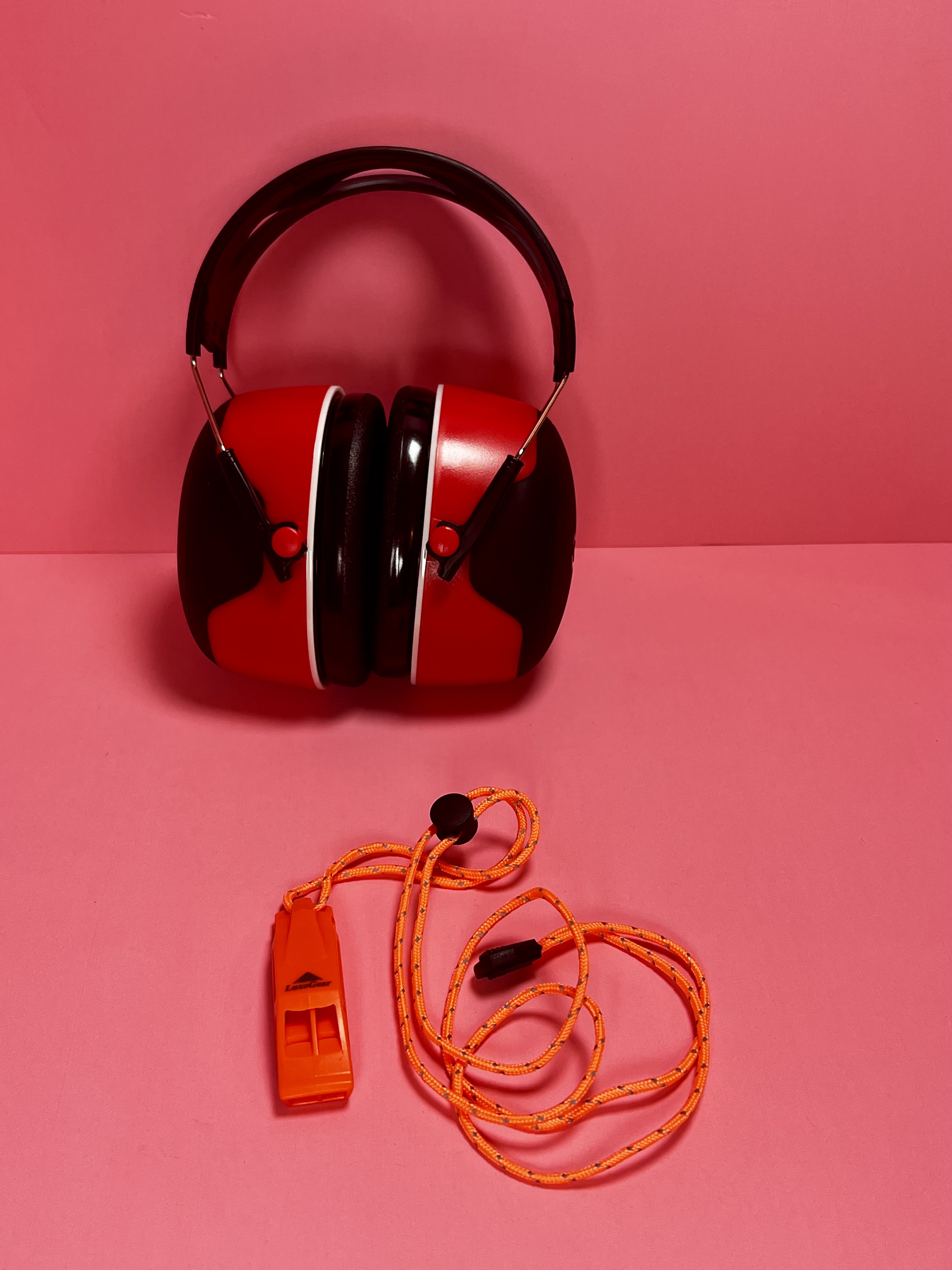 Orange emergency whistle placed beside red hearing protection earmuffs on a pink background.