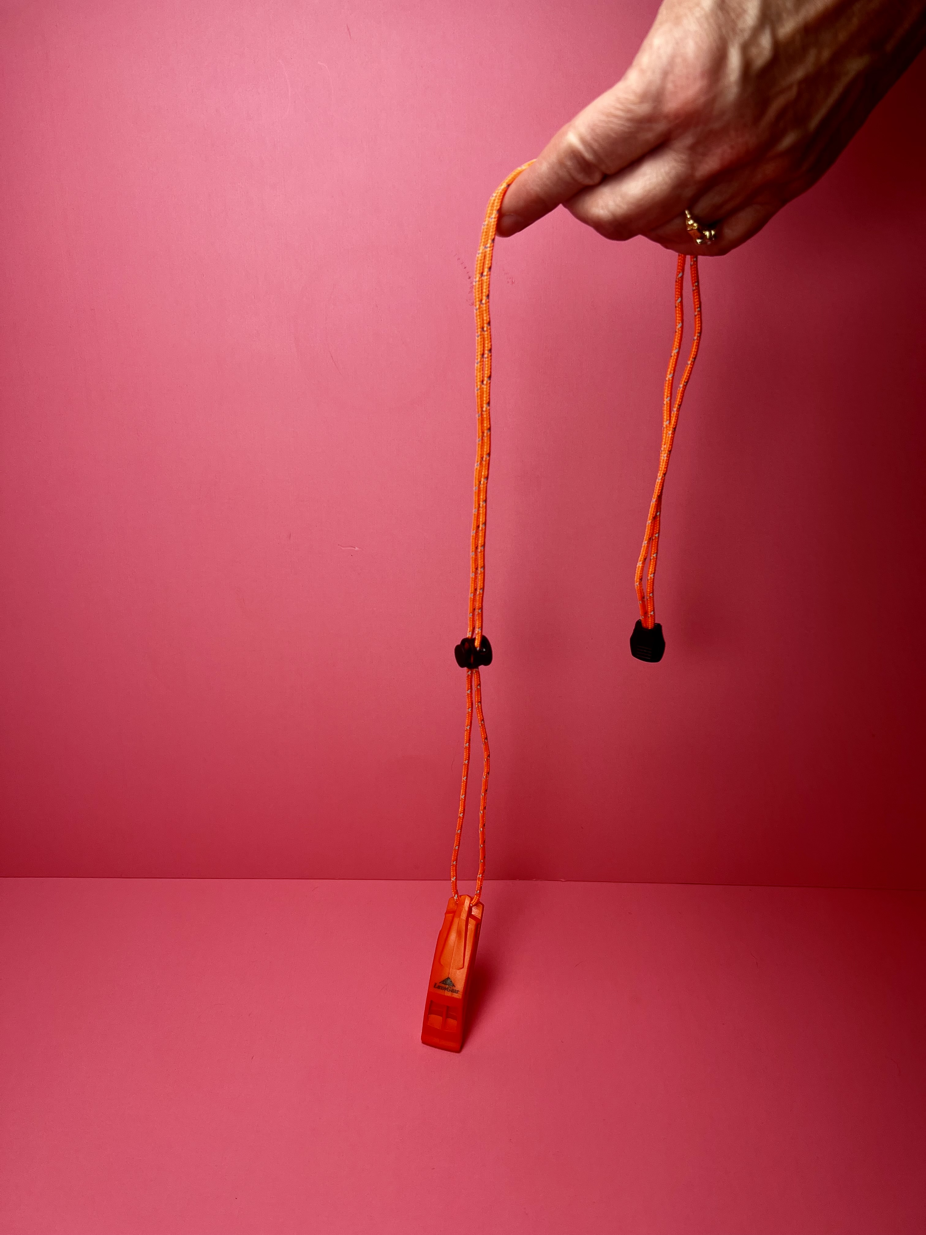 Hand holding an orange emergency whistle by its lanyard against a pink background.