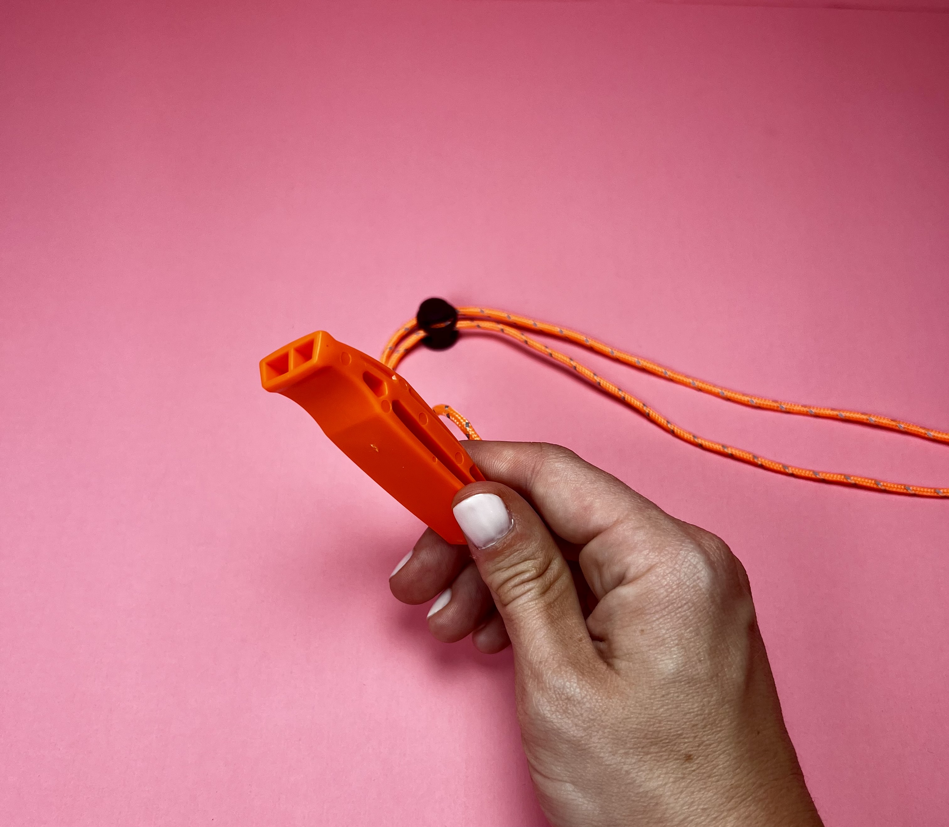 Close-up of a hand holding an orange emergency whistle with lanyard on a pink background.