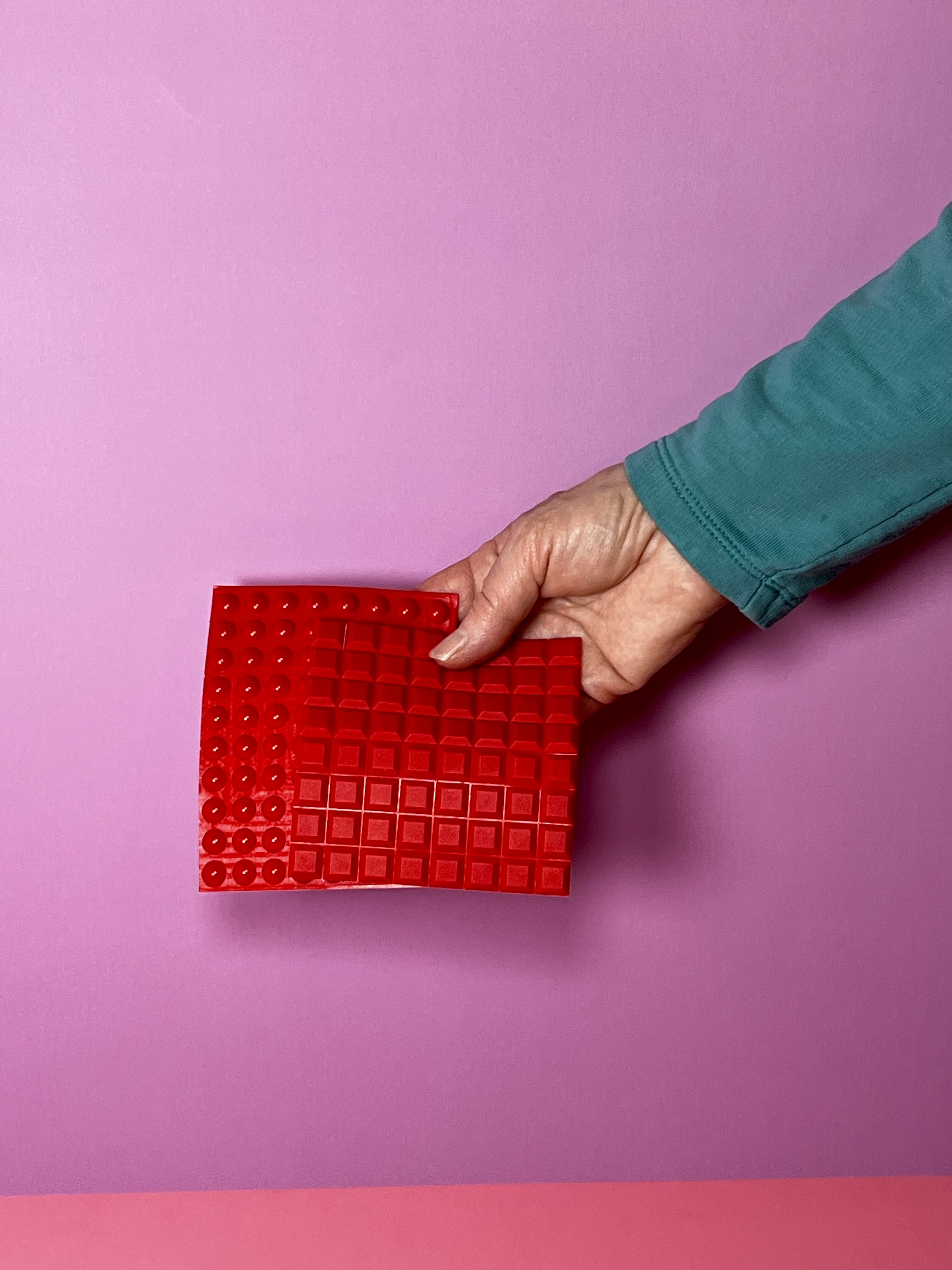 A hand holds a red sheet of raised tactile adhesive markers against a pink background.