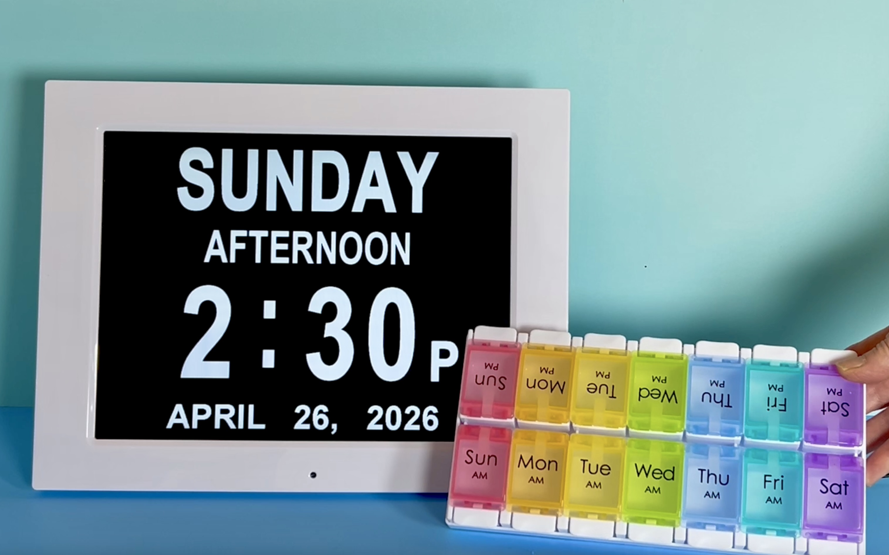 Digital day clock beside a colorful weekly pill organizer for medication management.