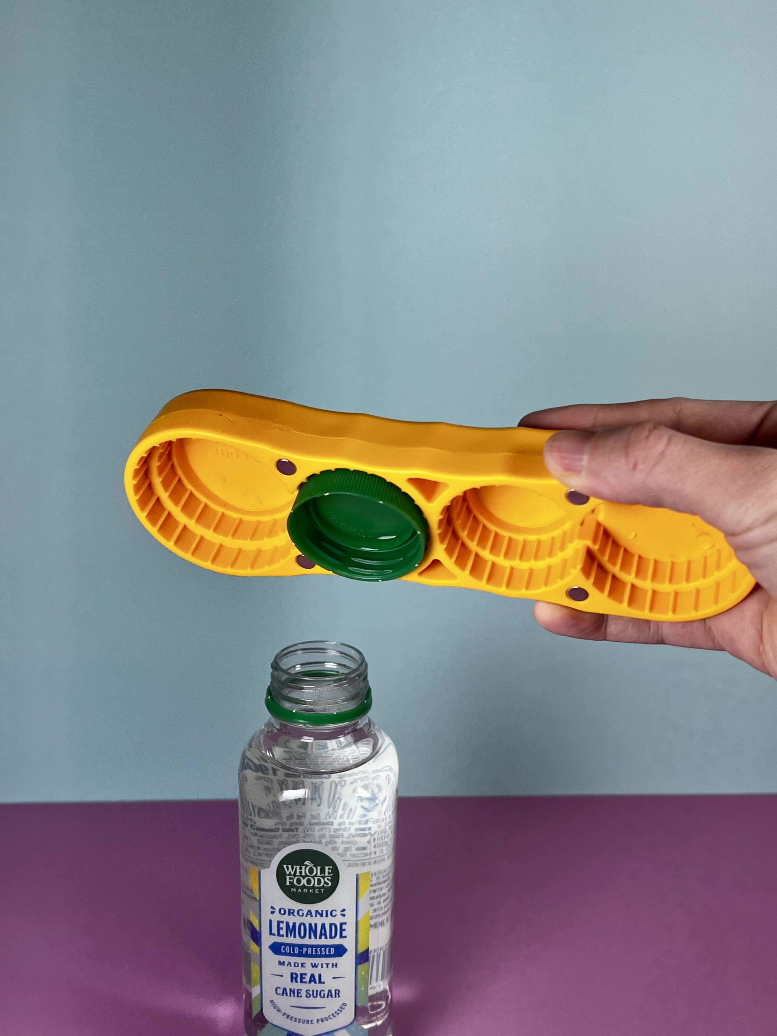 Hand using an orange multi-size bottle opener to remove a green bottle cap.
