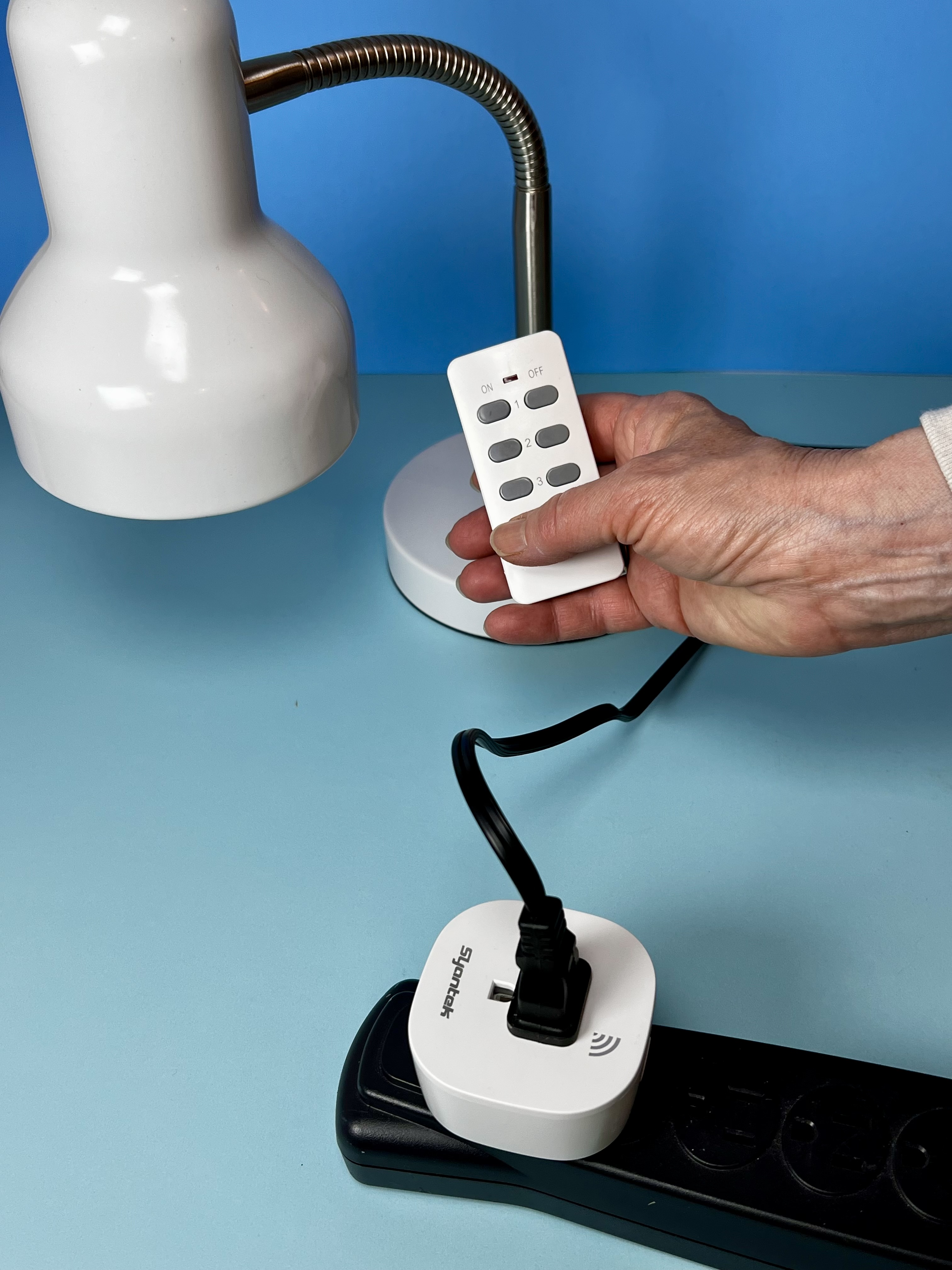 Hand holding a small white lamp remote beside a table lamp and connected plug.