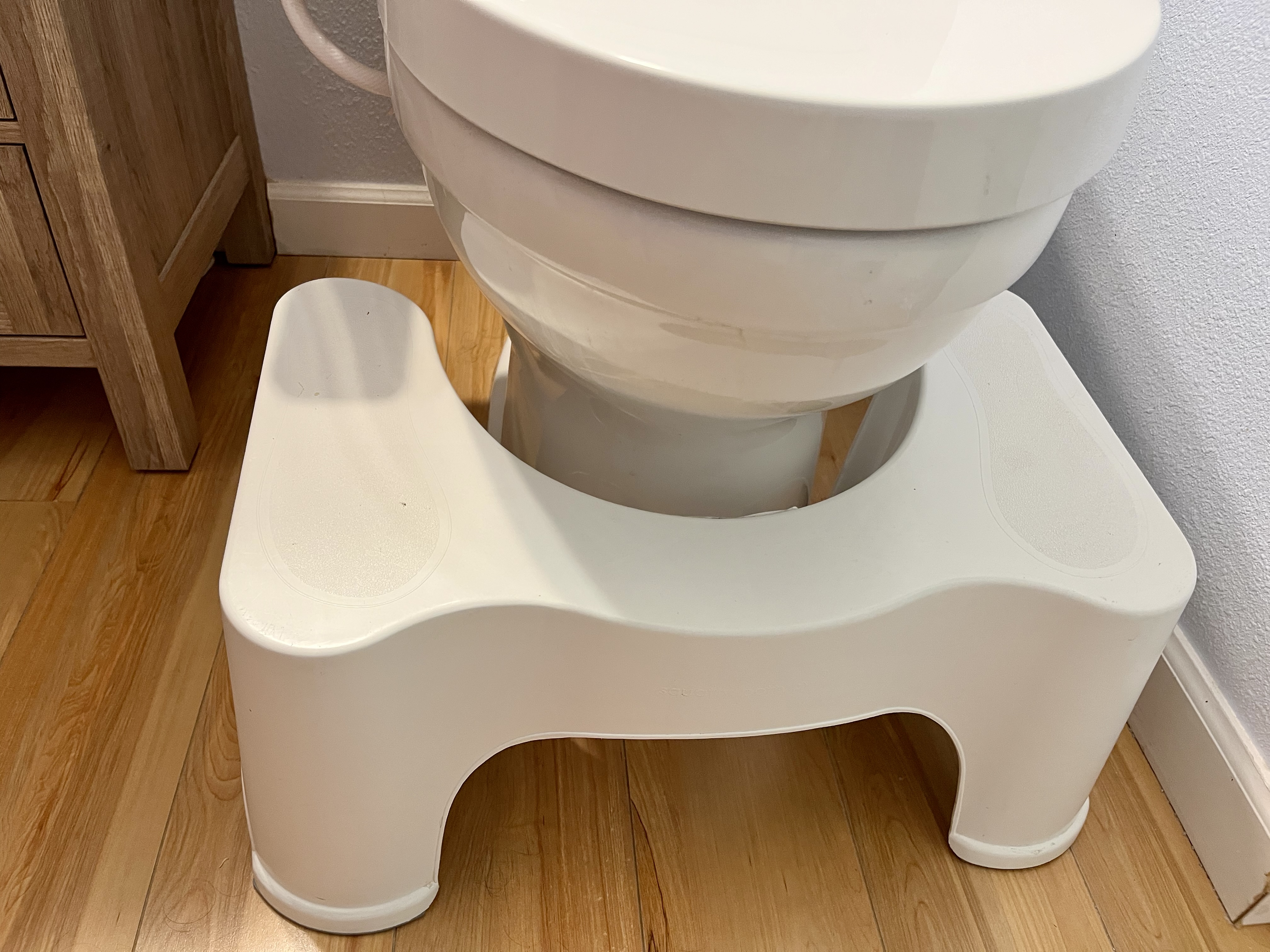 White toilet footstool positioned around the base of a toilet to raise the feet while seated.