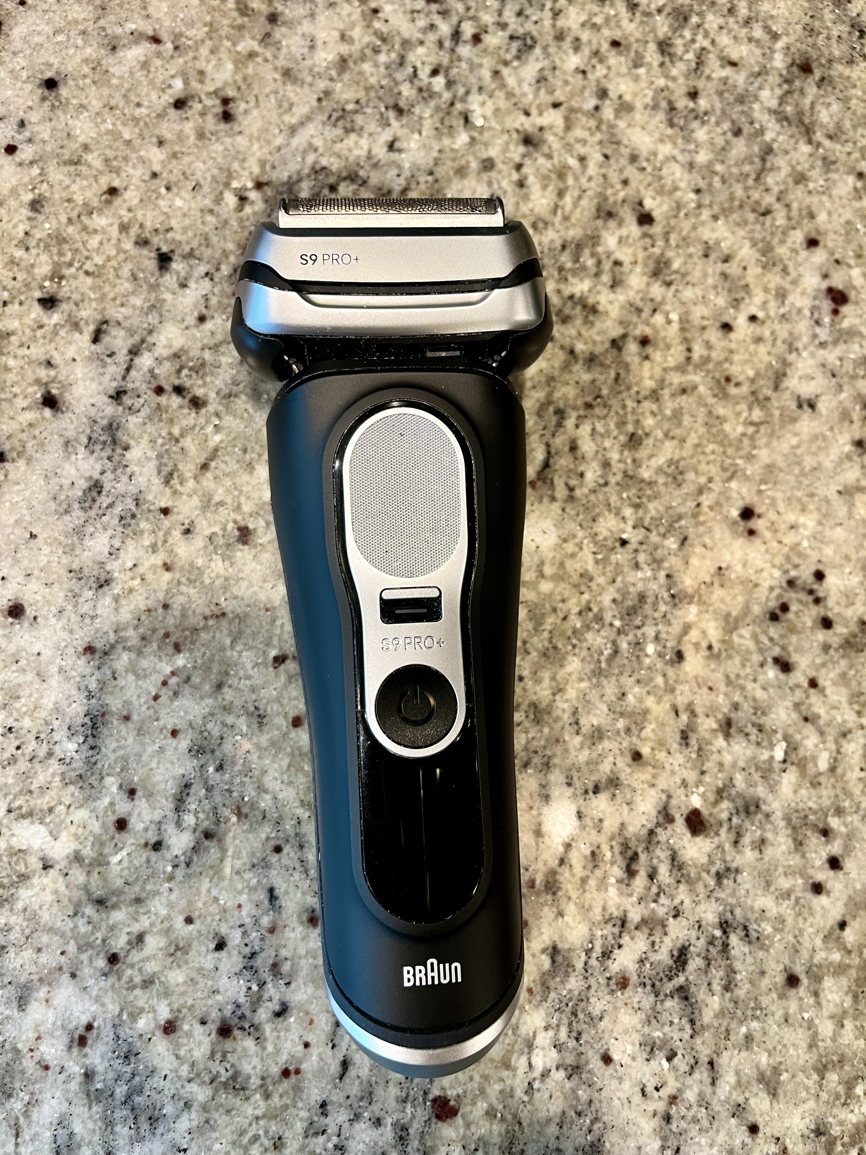 A black electric razor rests on a countertop, showing its large handle and shaving head.
