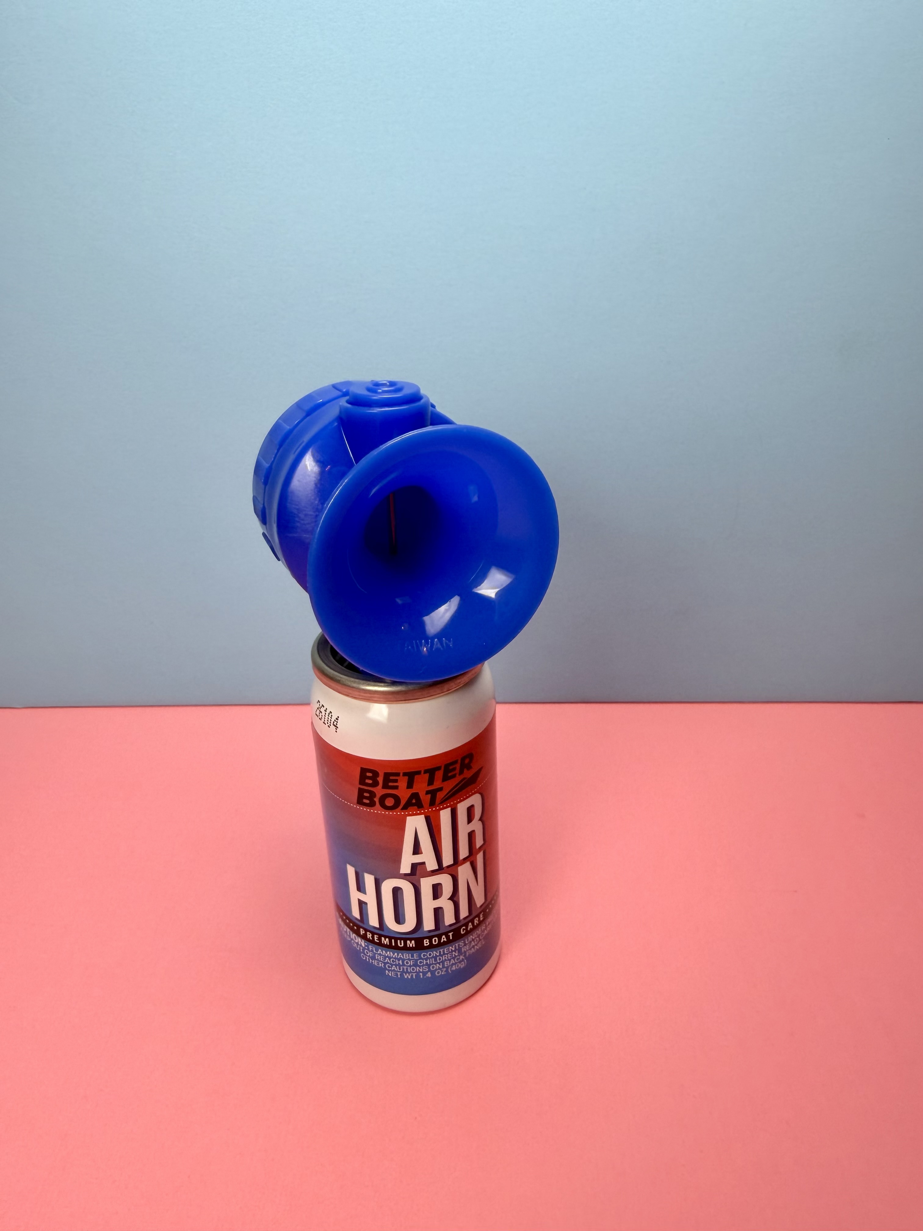 A small canister of Better Boat Air Horn with a bright blue trumpet horn top sitting upright against a pink and light blue background.