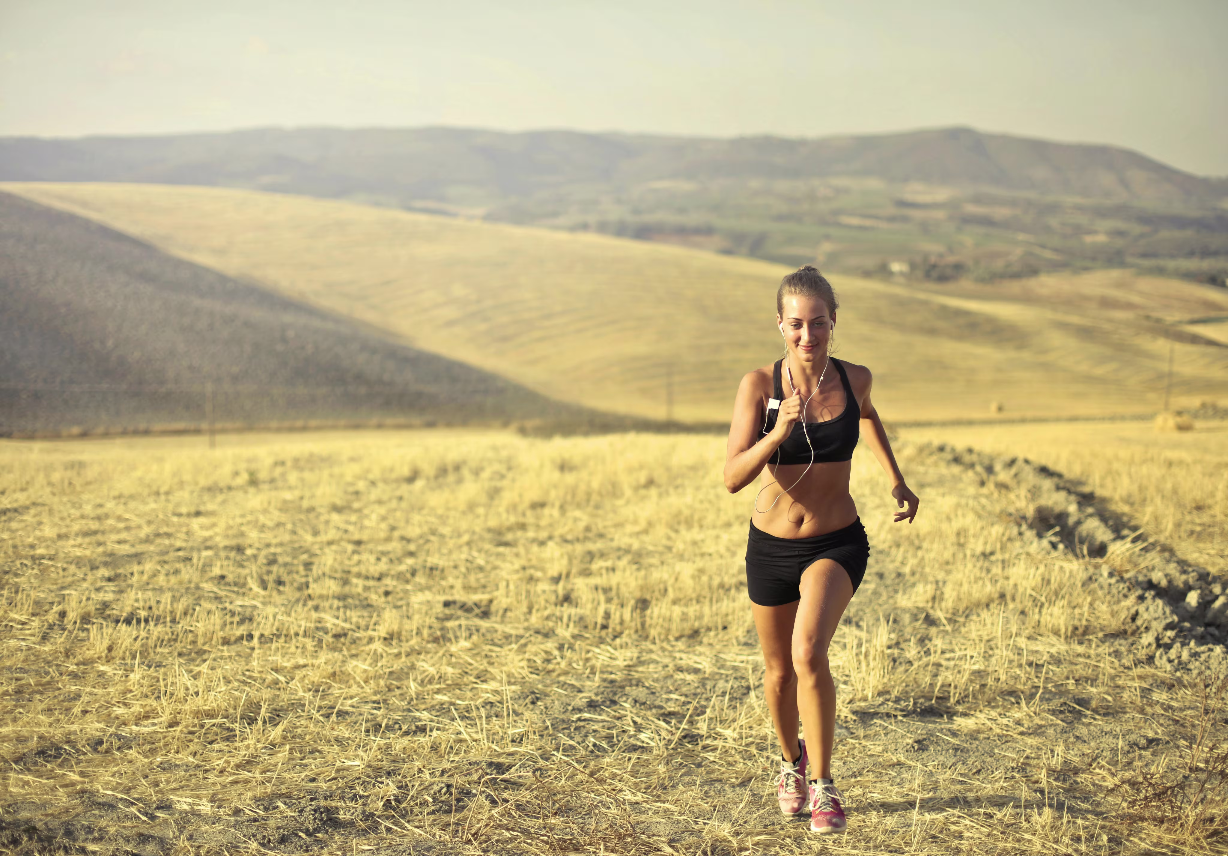Young woman jogging on a dry grassy field with hills in the background, wearing workout clothes and earphones.