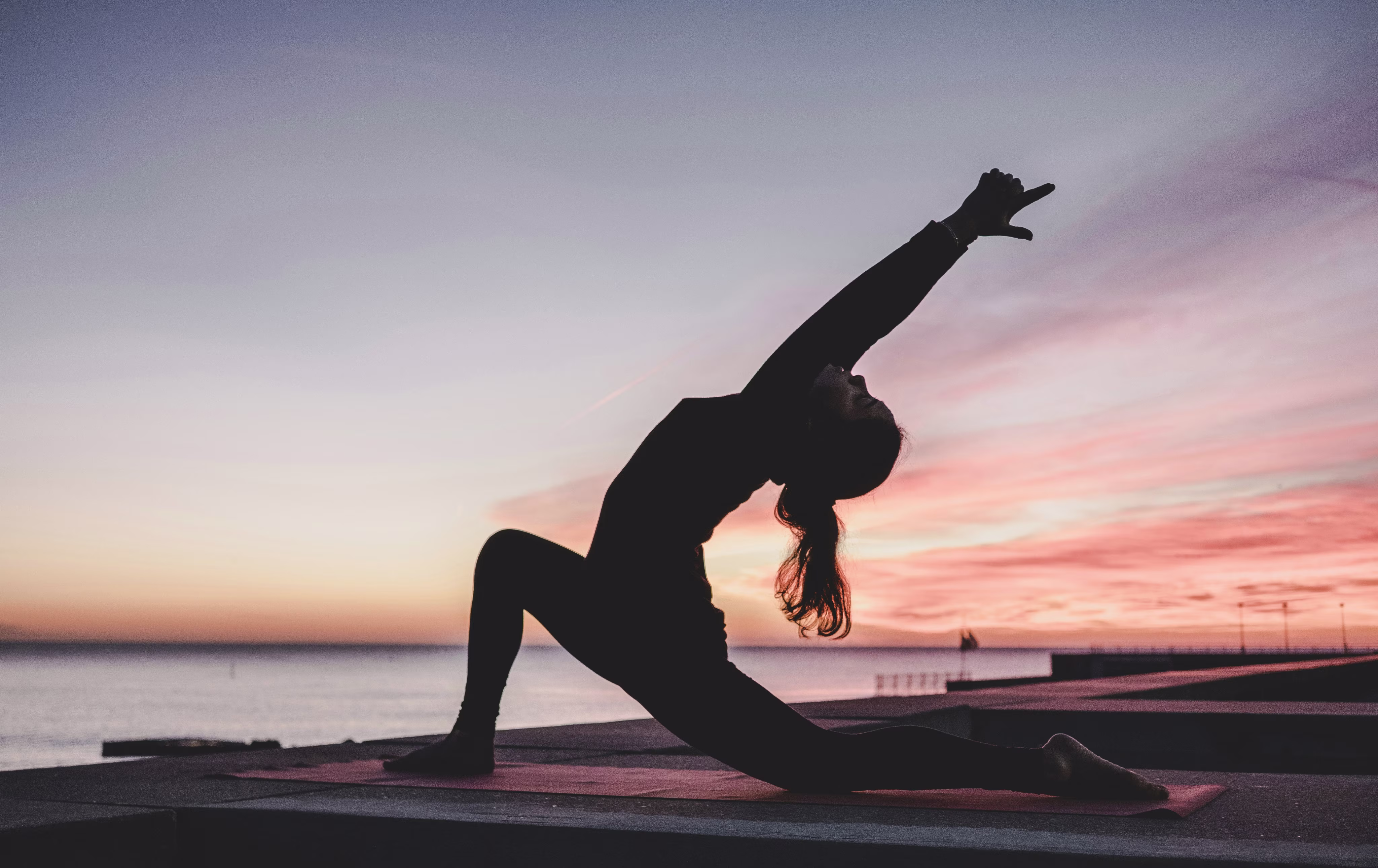 Silhouette of a person doing a yoga stretch on a mat near water during a colorful sunset.