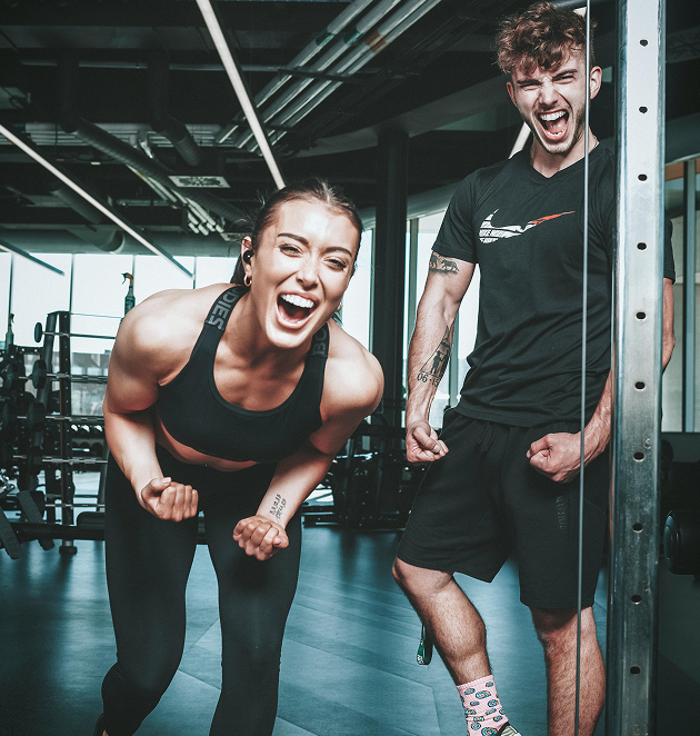 Two young adults in gym workout attire flexing muscles and smiling energetically.