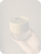 Hand holding clear glass cup filled with whipped coffee against a brown background.