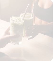 Two hands holding iced green smoothies with metal straws over a wooden table, one person in a black top.