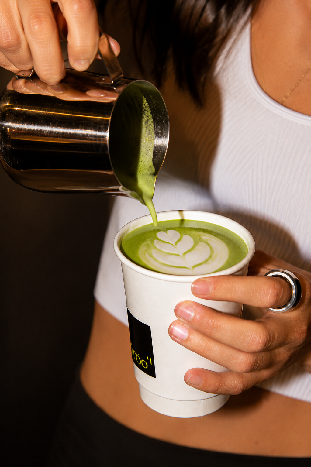 Person pouring green matcha latte with foam art into a white paper cup.