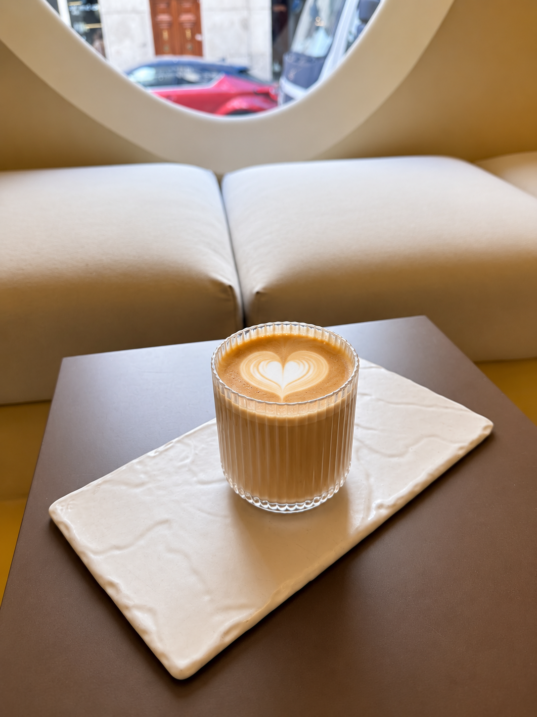 Glass of latte with heart-shaped foam art on a white tray on a brown table in a cozy café.