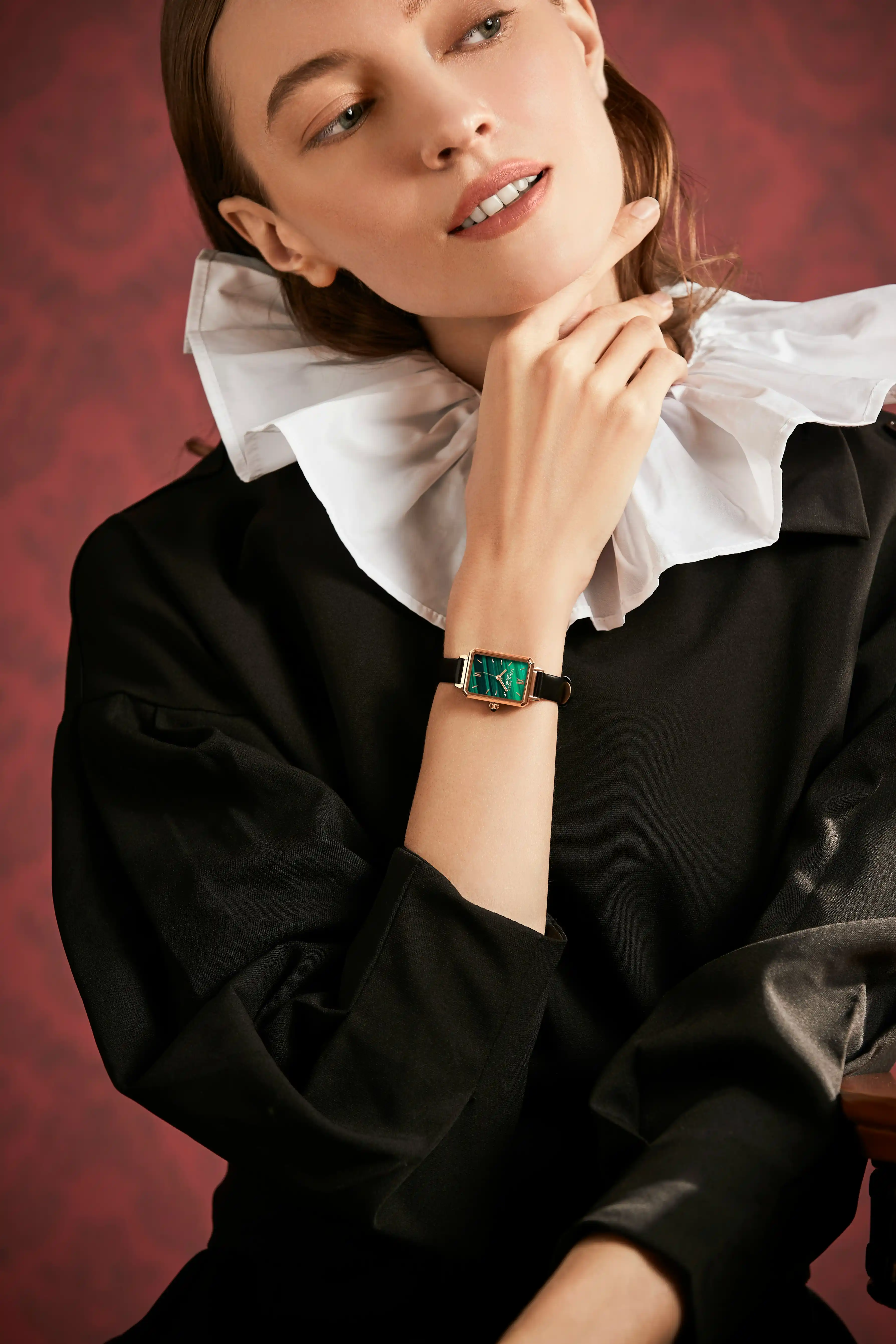 Woman in black dress with a large white ruffled collar wearing a rectangular green-watch with a black strap on her wrist, posing with her hand near her chin.