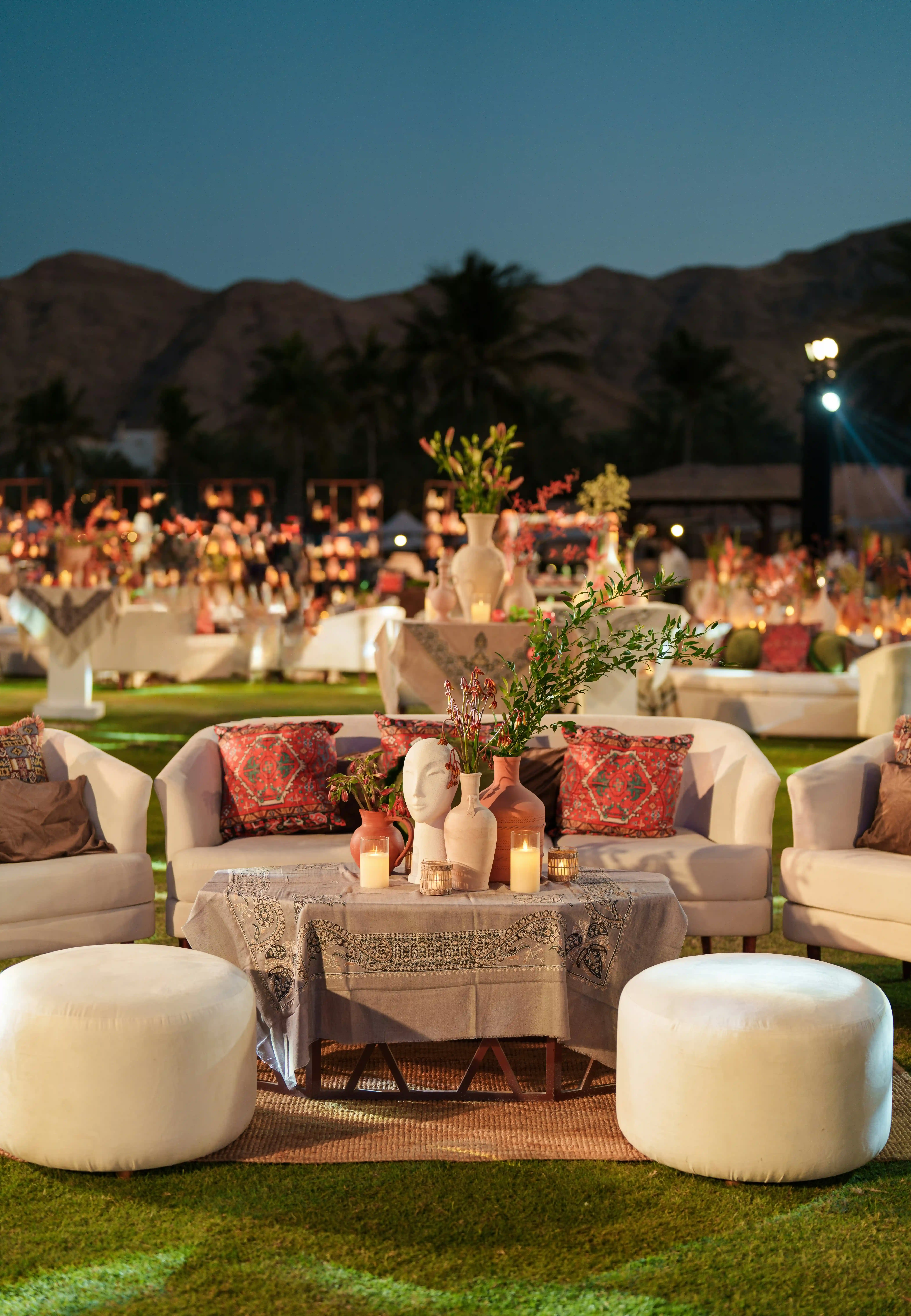 Outdoor evening seating area with white armchairs, round ottomans, a table covered with a patterned cloth, decorated with candles and vases containing greenery and a sculpted face.