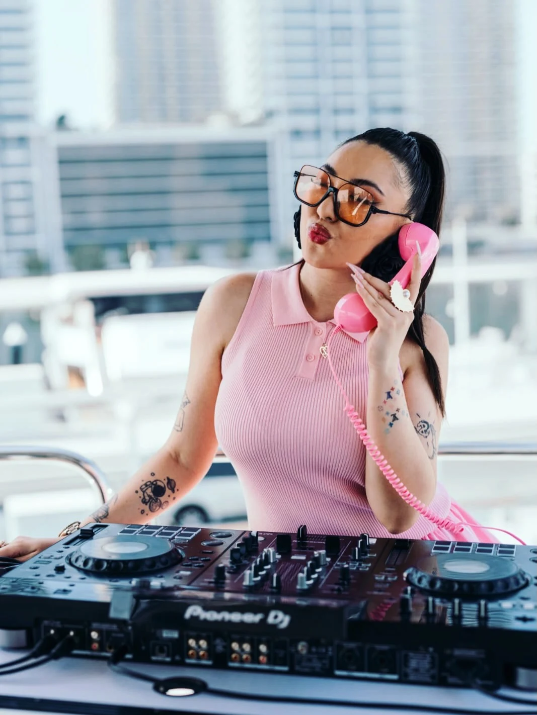 Female DJ in pink sleeveless top holding a pink retro telephone handset near her ear, with a DJ mixer in front of her and city buildings in the background.