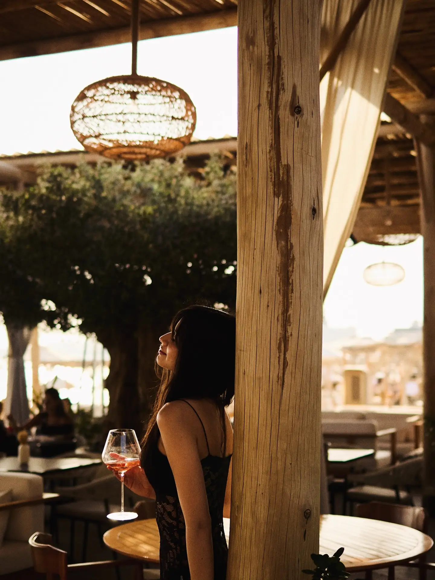 Woman in a black dress holding a glass of rosé wine, leaning against a wooden pillar in a warmly lit outdoor lounge.